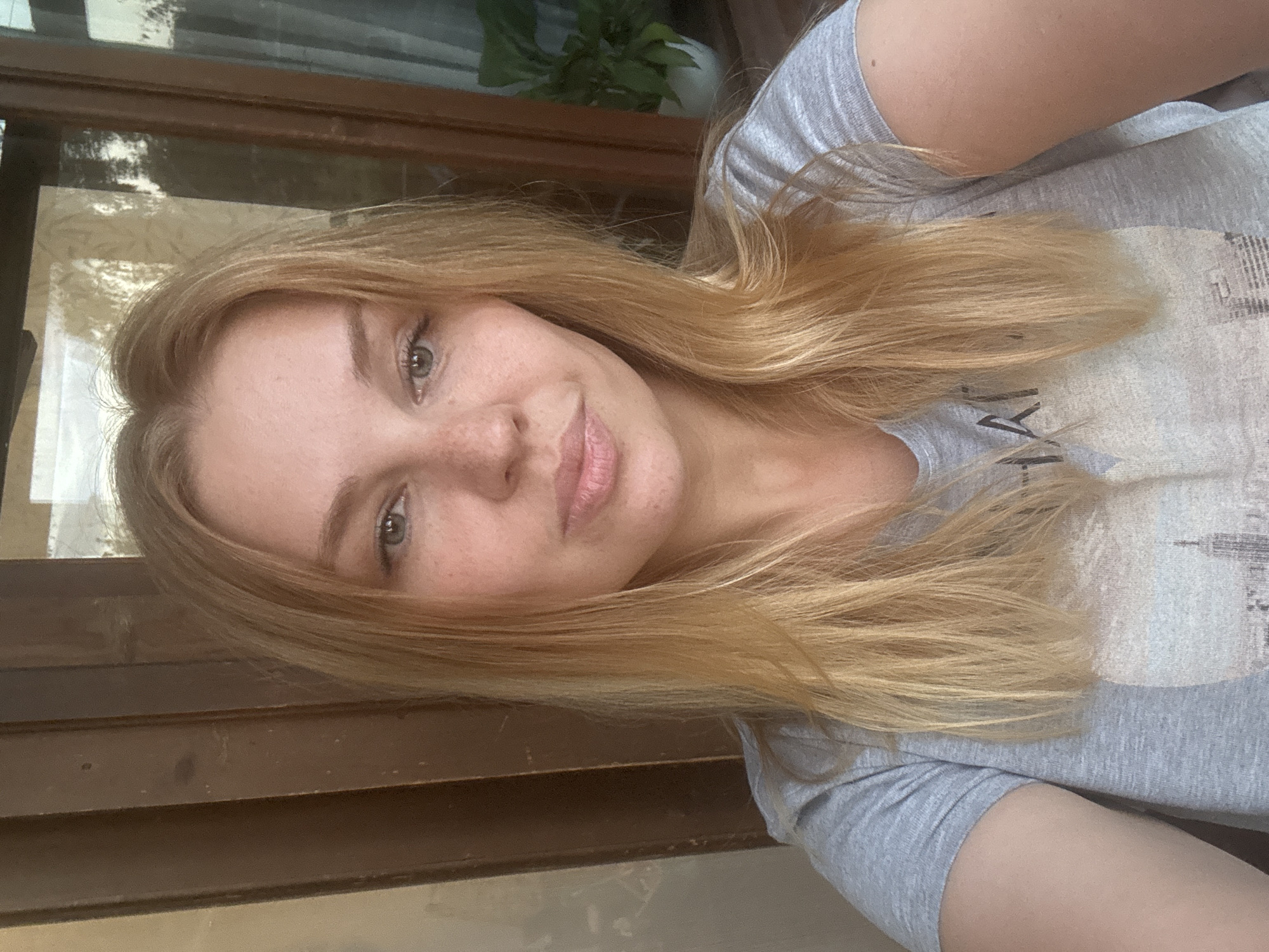 A young Caucasian woman with long blonde hair and freckles is pictured from the chest up. She is wearing a grey t-shirt with a printed image of a cityscape. She is looking directly at the camera with a neutral expression. The background consists of a wooden door and a window with reflections of trees and a potted plant. The setting appears to be outdoors, possibly on a porch or balcony, in Rusanovo, Russia.