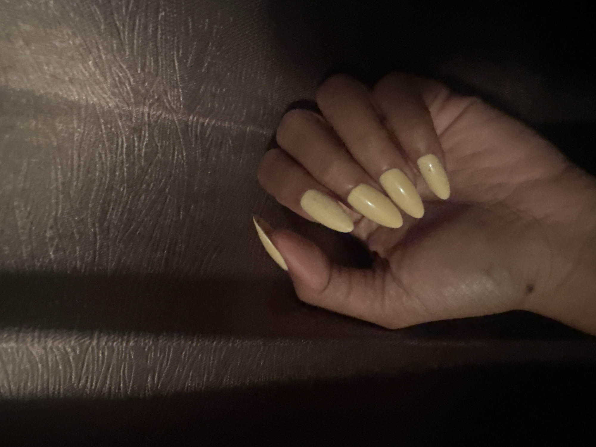 A left hand with long, almond-shaped fingernails painted a pale yellow is held up against a textured, dark brown background. The hand is the primary subject of the image, with the nails catching the light and appearing glossy. The background appears to be fabric, possibly a curtain or upholstery, with a ribbed texture. The lighting is dim, casting shadows and creating a moody atmosphere. There is no discernible location within Sukawati, Indonesia, provided by the image itself.