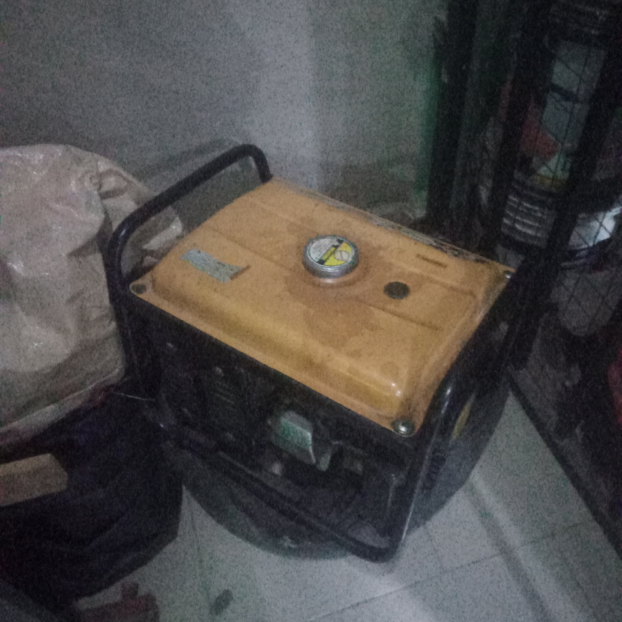 A yellow and black portable generator is visible in the foreground, situated on a tiled floor. The generator has a black metal frame around its top. A silver fuel cap with a yellow and black label is located on the top surface of the generator. To the left of the generator, a portion of a large, light-colored, possibly plastic-wrapped object is visible, and to the right, dark objects and shadows obscure details of the background. The walls in the background appear to be a mottled gray color. The scene is dimly lit.