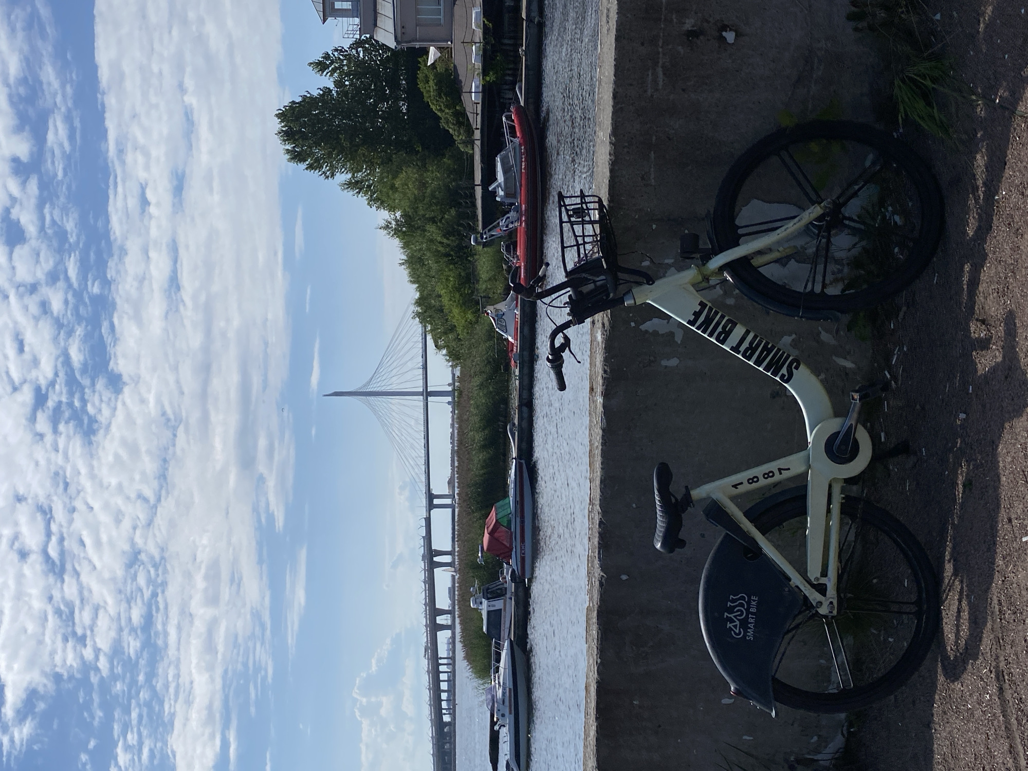 The image depicts a white and cream-colored bicycle with "SMART BIKE" branding on the frame and fender, parked in front of a concrete wall. The bicycle has a basket on the handlebars and the number "1887" is visible on the frame. In the background, several boats are docked on a body of water, with a large cable-stayed bridge spanning the water in the distance. Lush green trees and buildings are visible on the far bank. The sky is bright blue with scattered white clouds. This scene is consistent with a location along the Gulf of Finland in Saint Petersburg, Russia, possibly near the Western High-Speed Diameter bridge.