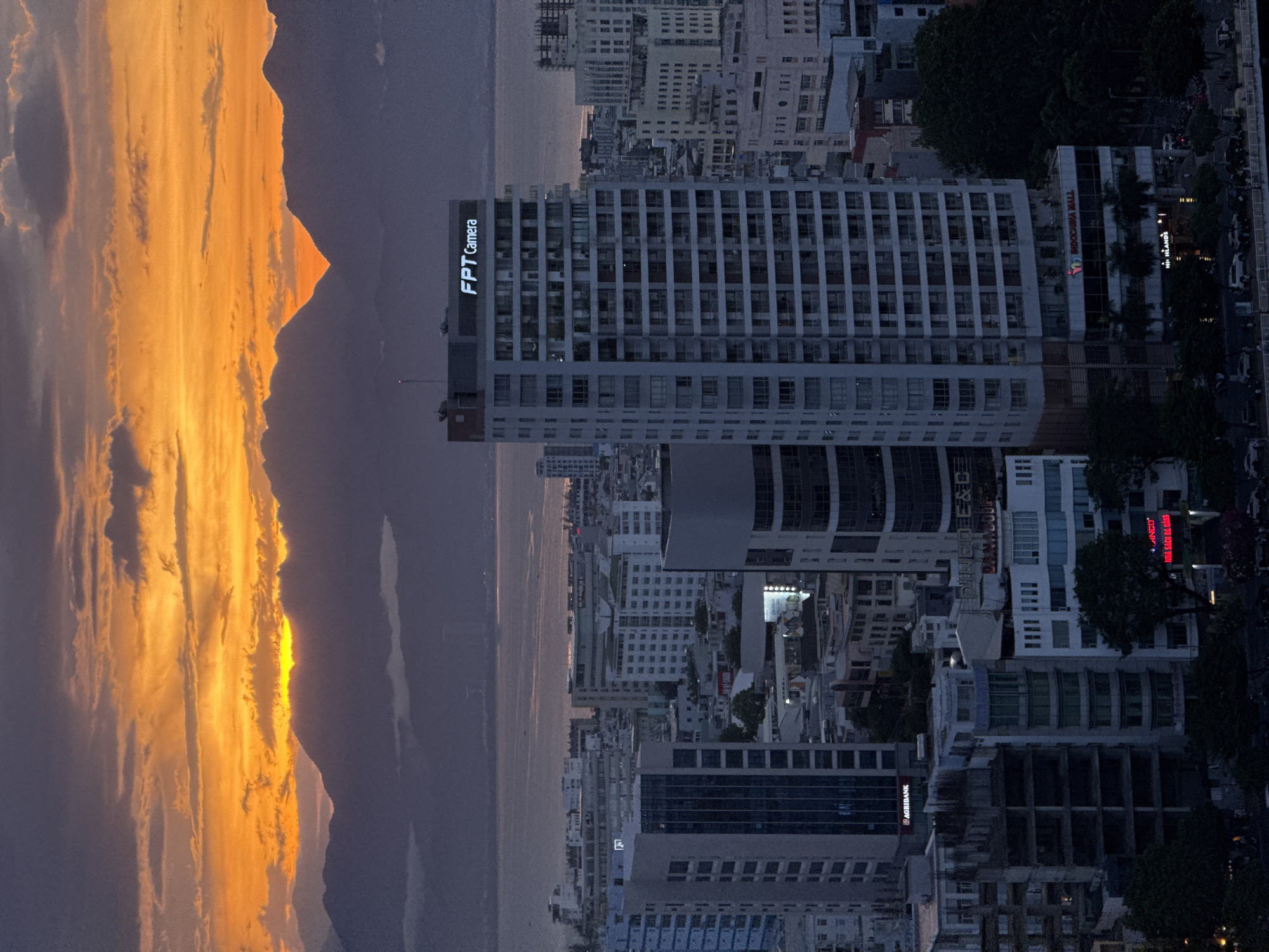 This is a high-angle, long shot of a cityscape at dusk in Da Nang, Vietnam. The sky is filled with vibrant orange and yellow clouds, illuminated by the setting sun. In the background, a range of mountains is silhouetted against the colorful sky, with a body of water visible between the mountains and the city.
The city is densely populated with numerous buildings, including several tall, modern high-rises. One prominent building in the center of the frame is a tall, light-colored skyscraper with many windows. The top of this building displays a glowing sign that reads "FPT Camera". Another building to its right, also a high-rise, is partially visible. In the foreground and midground, there are a variety of other buildings, some appearing to be under construction or in a state of disrepair, with exposed frameworks. Some buildings have illuminated signs, such as "AGRIBANK" and "INDOCHNA MALL". Trees and dark foliage are visible in some areas of the city. The streets are visible at the bottom of the image, with several cars and what appear to be motorcycles parked or moving. The overall scene captures the transition from day to night in a bustling urban environment with a dramatic natural backdrop.
