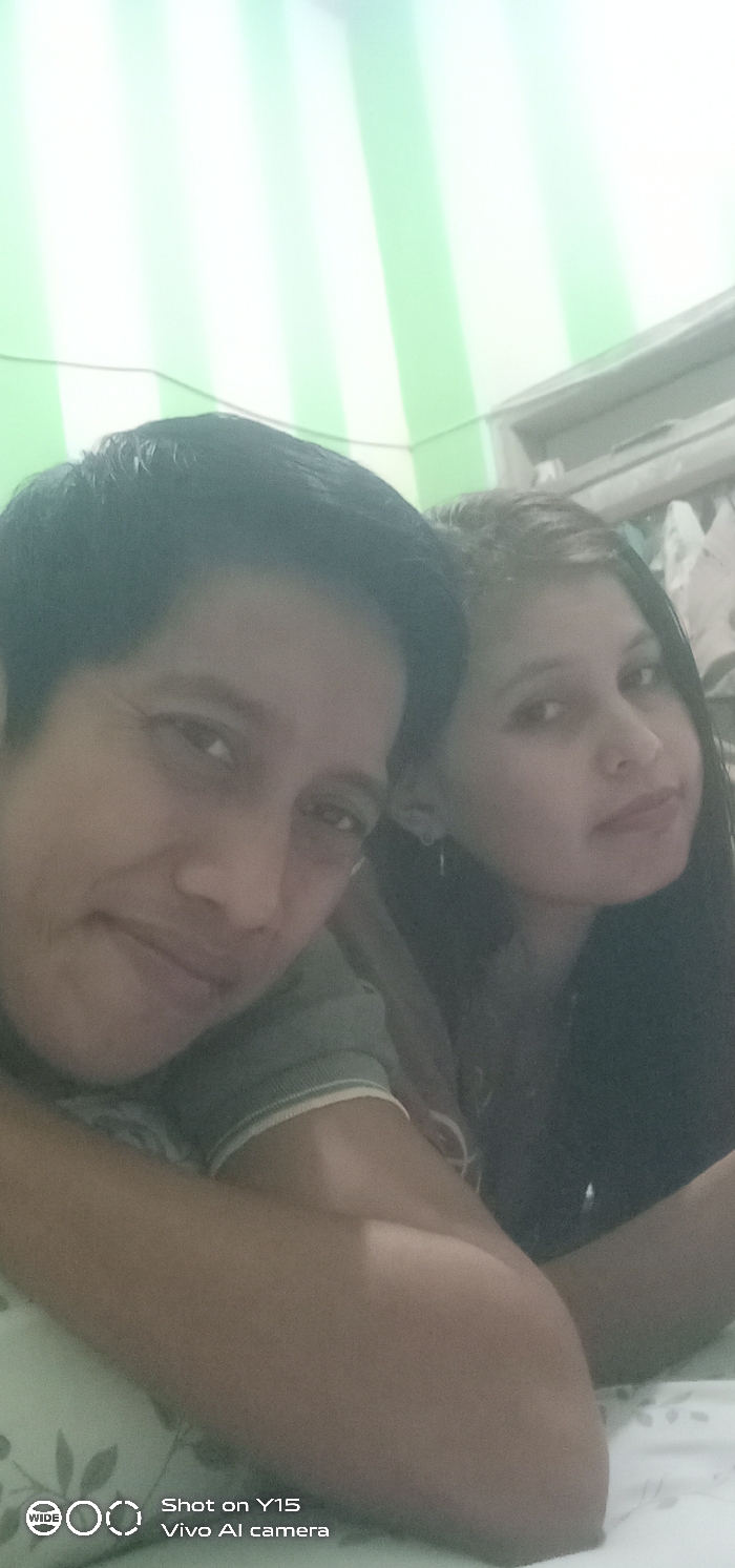 This image, captured in the city of Ranugedang, Indonesia, depicts two adults in a close-up, indoor setting. The individual on the left, identified as male, has short dark hair and is wearing a green short-sleeved t-shirt with a light-colored ribbed collar. He looks towards the camera with a slight smile, his left arm visible in the foreground, bent at the elbow and resting on a white fabric with a subtle light green leaf pattern. The individual on the right, identified as female, has long dark hair and is wearing a dark-colored top; she also looks towards the camera with a neutral expression. They are positioned in close proximity, appearing to be posing for a photograph. The background features a wall with vertical stripes of light green and white. To the right, a portion of a light-colored frame, possibly a window or door, is visible, with indistinct objects behind it. The image contains a "Shot on Y15 Vivo AI camera" watermark in the bottom left corner.
