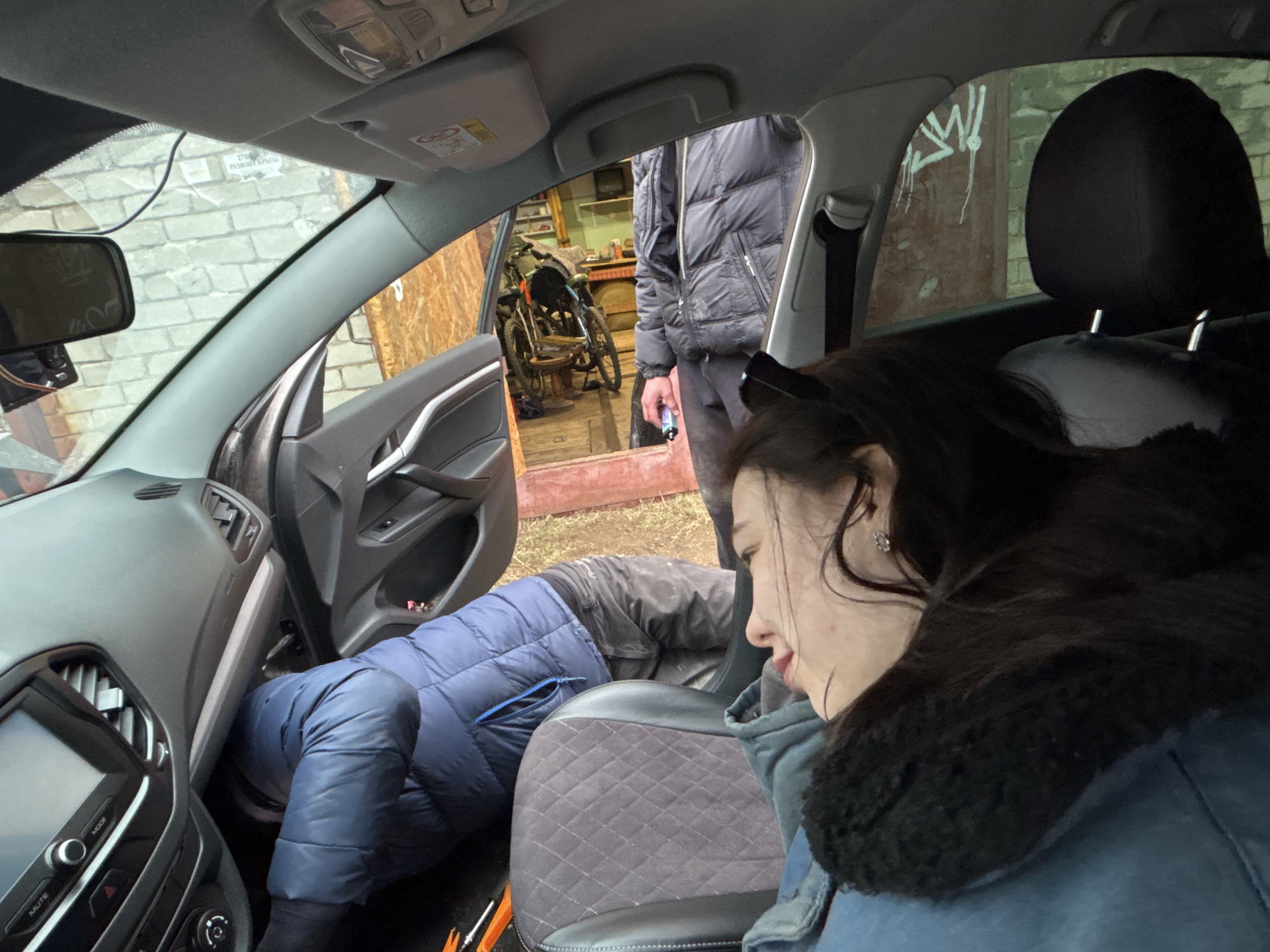 A medium shot from inside a car, likely in Perm, Russia, shows a woman sitting in the passenger seat while a man in a blue puffer jacket is bent over, apparently working on something under the dashboard of the driver's side. Another person stands outside the open driver's side door, partially visible in a black puffer jacket. The car is parked in front of a garage with bicycles and items inside. Graffiti is visible on the exterior wall of the garage. The interior of the car is modern.