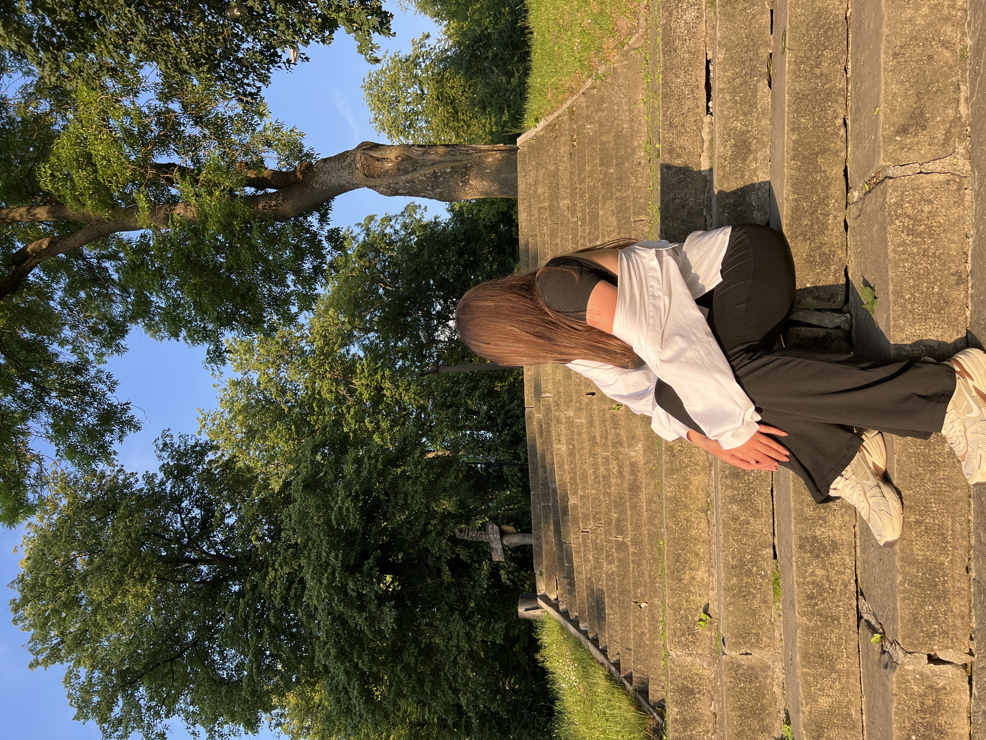 The image displays an outdoor scene during the golden hour, characterized by warm, soft sunlight and long shadows. A young person, seen from a slightly elevated angle and from behind, is seated on old, weathered stone stairs. The individual wears a white long-sleeved shirt over a dark top, black wide-leg pants, and light-colored sneakers. Their long, straight brown hair cascades forward, obscuring their face as they sit with their head bowed, suggesting a moment of rest or contemplation.
The stone stairs, likely made of concrete or a similar material, show signs of age, with visible cracks and small plants growing in the crevices. They descend through a natural setting, flanked by vibrant green grass and dense foliage. Numerous large, mature trees with lush green leaves dominate the background and left side of the frame, their branches stretching against a clear, bright blue sky. One prominent tree trunk is visible, adding to the natural grandeur. In the mid-ground, nestled among the trees and grass near the stairs, a stone structure resembling a cross or monument is partially visible, hinting at a historical or sacred context. The absence of specific activity beyond sitting creates a tranquil and serene atmosphere. There is no visible text in the image.
The setting, with its old stone structures and abundant natural greenery bathed in warm light, aligns with a location such as Zbarazh, Ukraine, which is known for its historic sites and picturesque landscapes.