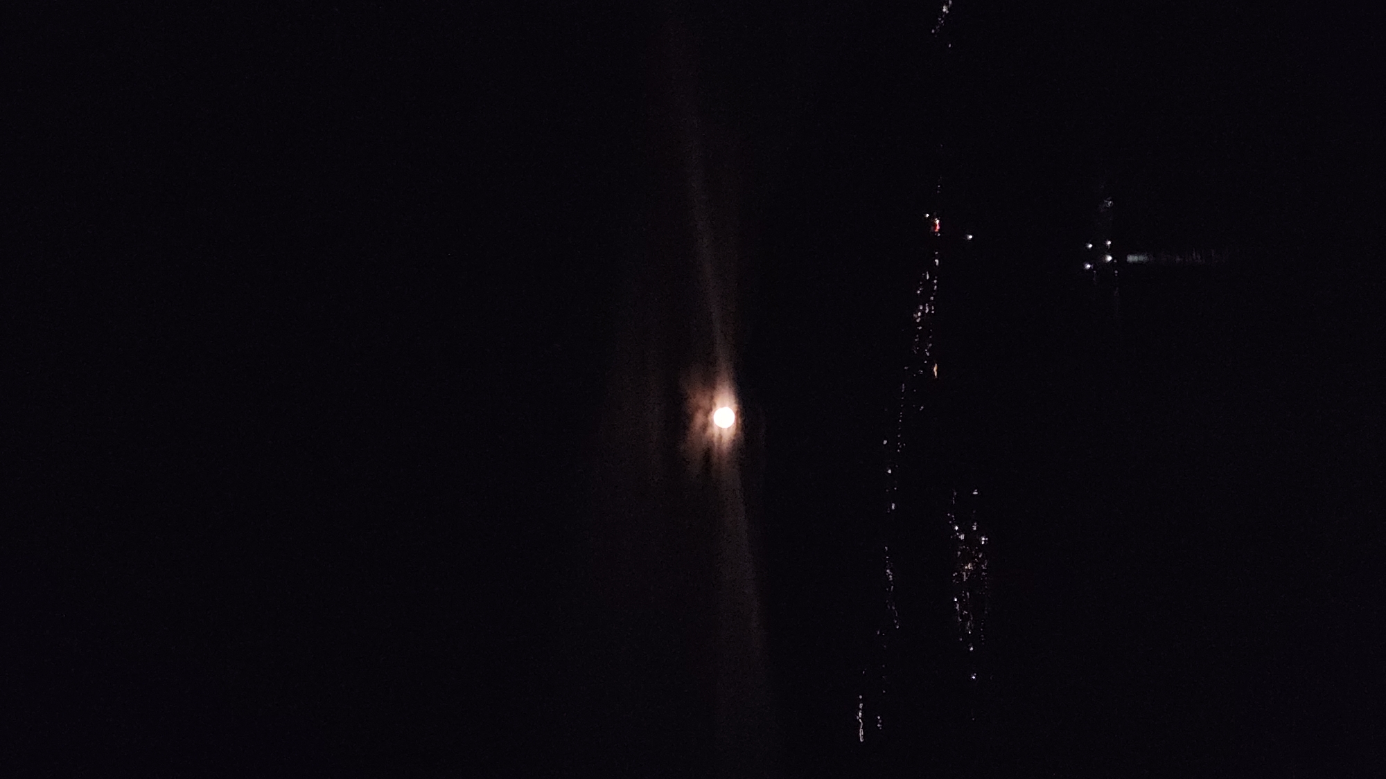 A low-light photograph shows the moon over water, reflecting its light. To the right of the moon's reflection, a faint line of city lights is visible. Given the location of Budva, Montenegro, it is likely that the photograph was taken from the coast, with the moon reflecting on the Adriatic Sea. The specific details in the cityscape, however, are difficult to discern due to the darkness.