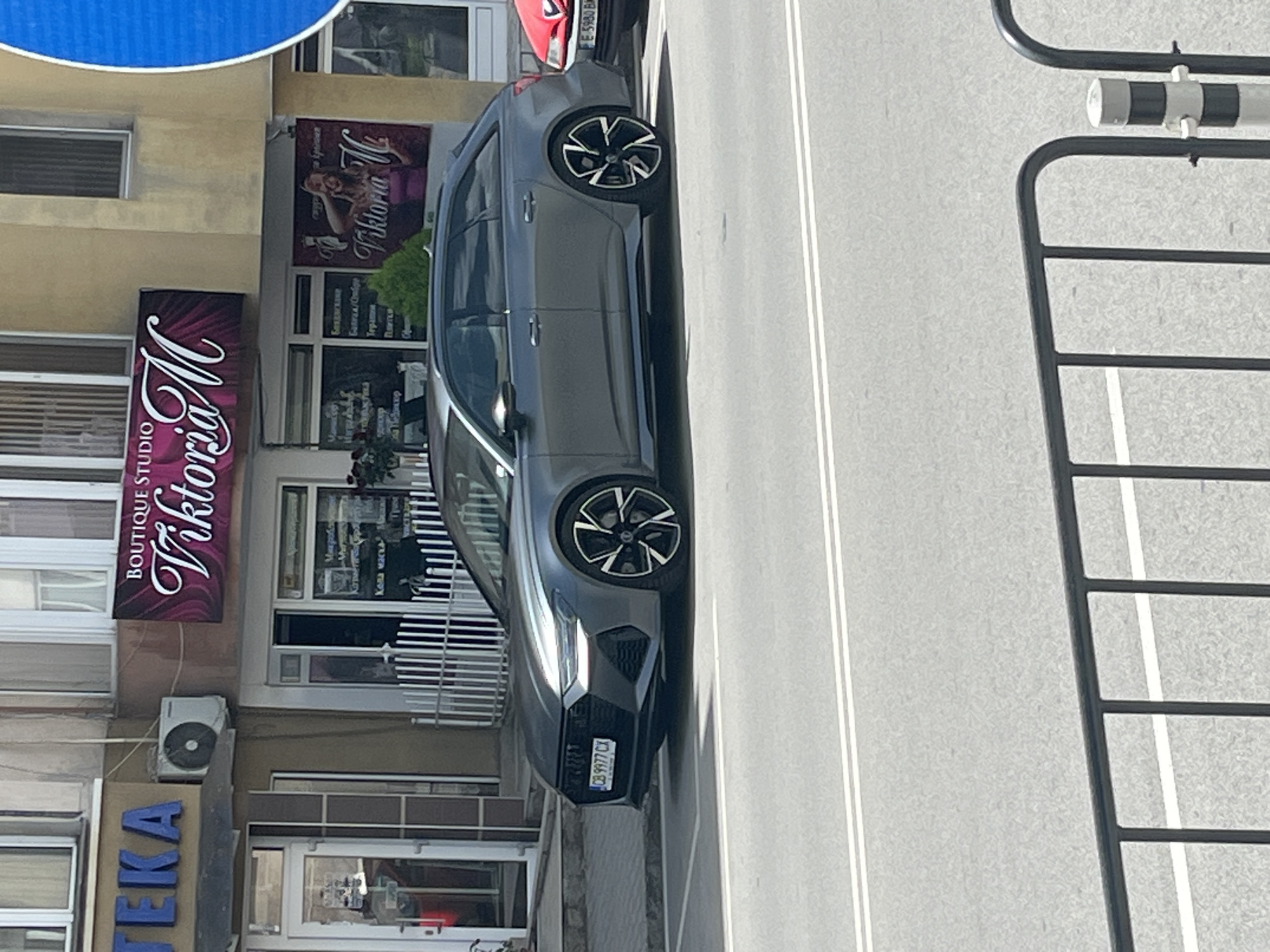 The image shows a gray Audi A7 parked on a street in an urban area. The car has a Bulgarian license plate, CB 9977 CX. To the left of the car, storefronts display signs for "Viktoria M Boutique Studio" and "LEKA" (pharmacy or drugstore). Further up is a sign with "Viktoria" written in cursive with an image of a woman next to it. There is also a sign containing text in Bulgarian. The street is marked with white lane lines. A black metal fence is visible on the right side of the image, likely part of a barrier or traffic control device.