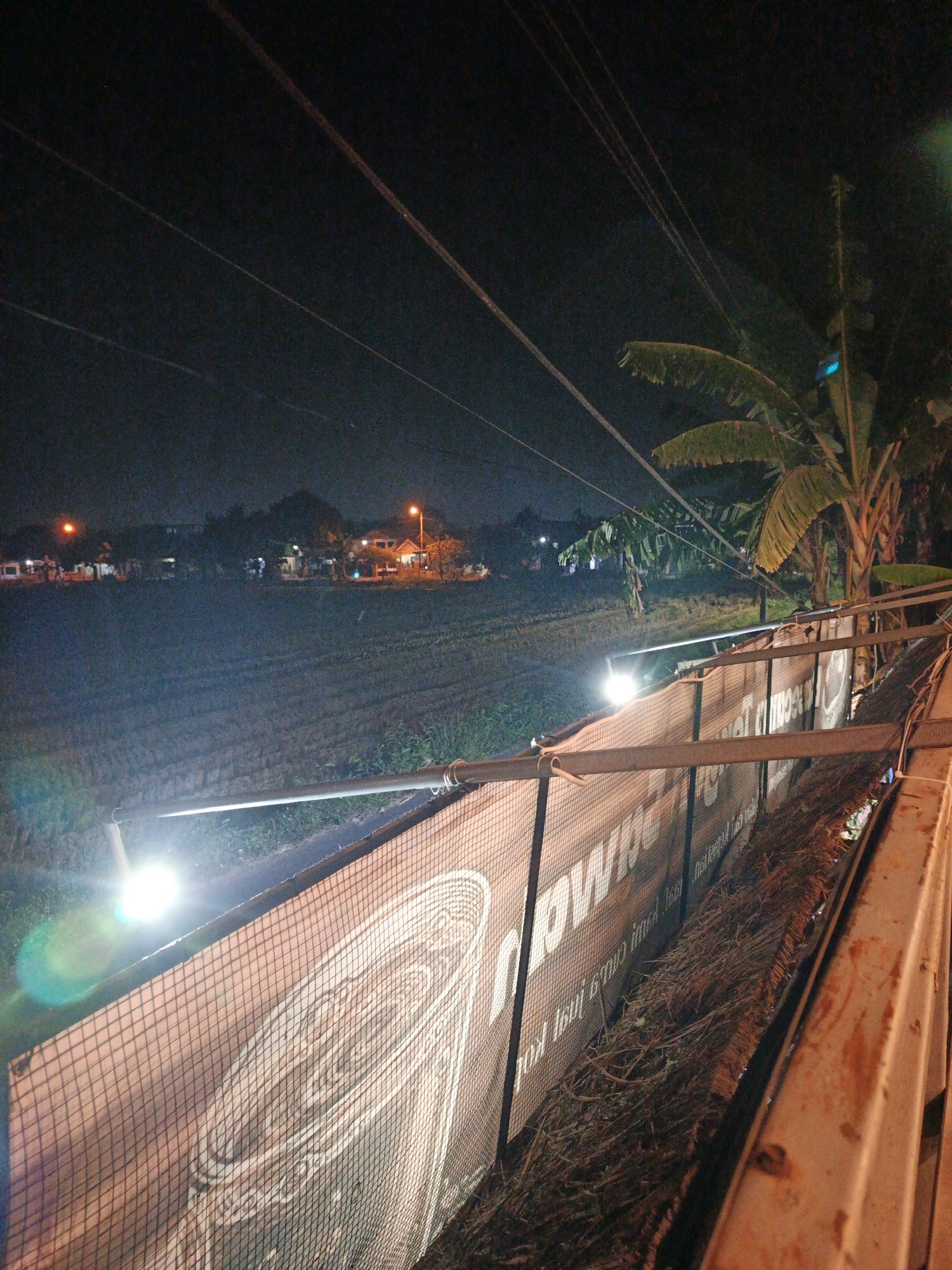 An exterior, eye-level, night shot captures a fence adorned with a banner in the foreground, a field with lights in the midground, and a residential area with lights in the background, all under a dark, clear sky in Ketegan, Indonesia. The fence, constructed from metal poles and mesh, is partially covered by a large banner displaying white lettering and a spiral design, illuminated by a bright light on the left side. The field, with furrows, is lit by several bright lights. A cluster of houses with glowing windows and streetlights is visible in the distance, partially obscured by trees, including a large banana tree on the right. Overhead, thin wires crisscross the sky. No people are visible in the scene.
