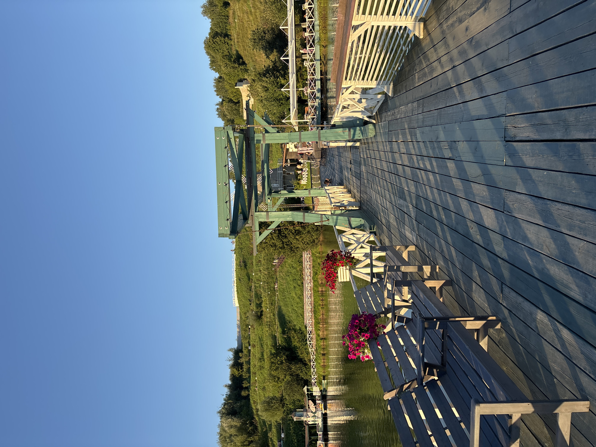 The image depicts a wooden pier extending over a body of water. The pier is made of dark wooden planks and is lined with white railings. Several wooden benches are positioned along the left side of the pier, some with potted pink and red flowers. In the background, a lush green hill rises, dotted with trees and some structures. A white pedestrian bridge crosses the water in the mid-ground. On the water, a small boat or ferry is docked. The sky is a clear, bright blue. The scene appears to be in the city of Romashkovo, Russia, and the lighting suggests it is late afternoon or early evening. A few people are visible in the background, scattered along the hill and on a covered seating area across the water. The overall atmosphere is serene and picturesque.