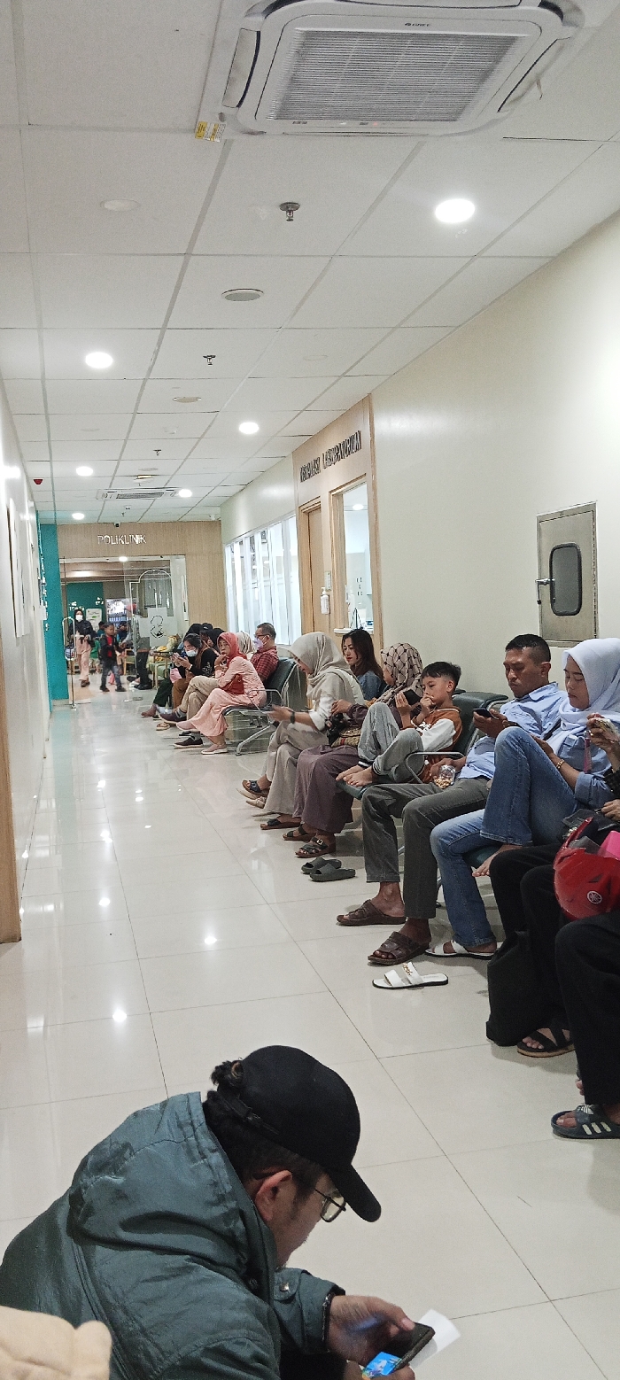 The image depicts a waiting area in a clinic, located at the coordinates -7.032 latitude and 107.527 longitude in Soreang, Indonesia. The area is lined with white tiled walls and floors, with a white ceiling containing several recessed lighting fixtures and a single air conditioning unit. The room contains a group of people, both adults and a child, all seated on a line of gray cushioned chairs facing the wall. Some individuals are looking down at mobile devices, while others are engaged in conversation. The clinic's name, "POLIKLINIK," can be seen written on a wall to the left, along with a closed door. To the right of the chairs are two closed doors marked "Klinik Gigi Umum." The rightmost door is a glass-panelled door, and the door on the left is wooden with a "no entry" sign attached.  A man seated on the far right of the image is wearing a blue shirt and jeans. A woman next to him is wearing a pink hijab, with a black bag in her lap. In front of them is a young boy wearing a gray shirt and jeans, holding a mobile device. Next to him is a woman wearing a white hijab.  The image is focused on a man sitting on the floor, with his back to the camera, facing the group of seated individuals. He is wearing a black baseball cap, a dark green jacket, and black pants, and he is looking down at his phone.