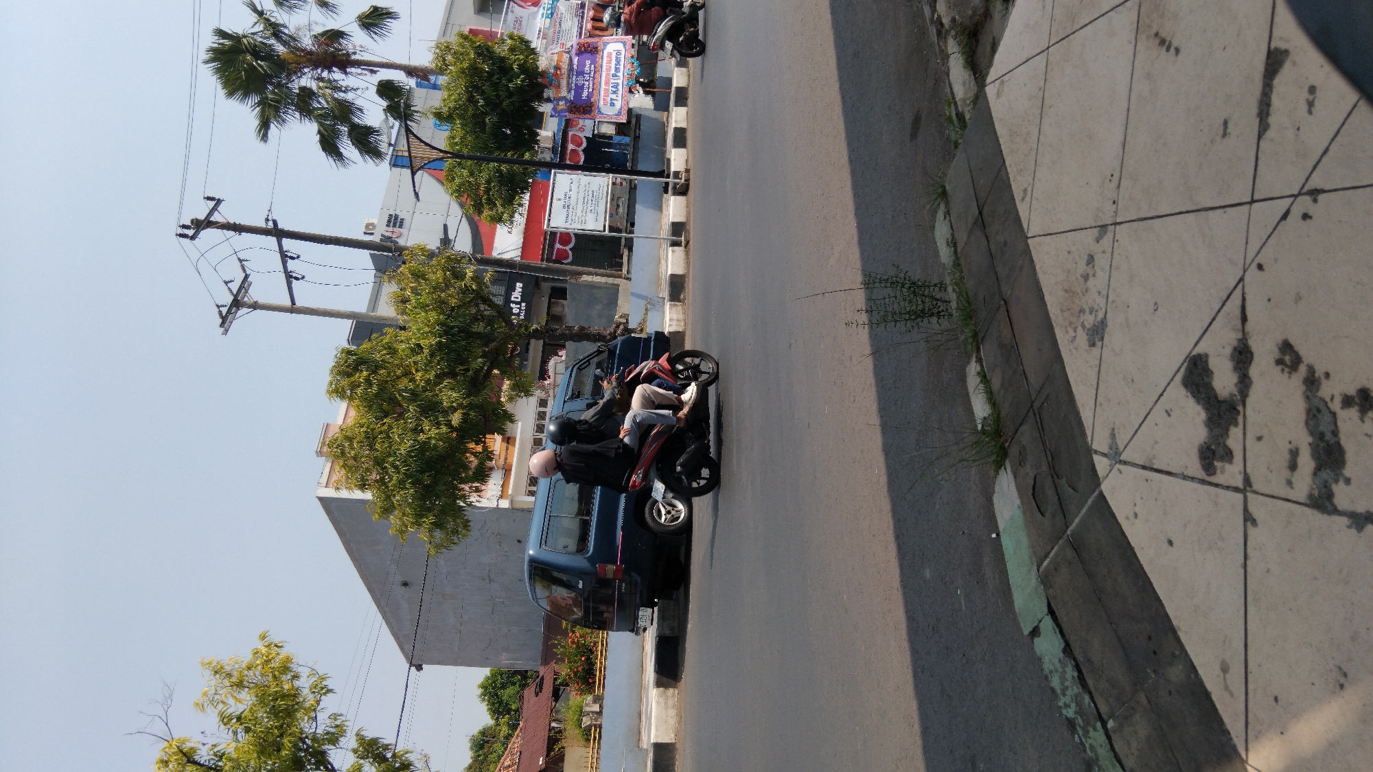 Two individuals are riding a red motorcycle on a paved road in Batang, Indonesia. The person in the front is driving, while the person in the back is a passenger. Both are wearing helmets. A dark blue van is parked on the side of the road behind the motorcycle. In the background, there are buildings, trees, and utility poles. The sky is clear and blue. The ground in the foreground is a tiled sidewalk.