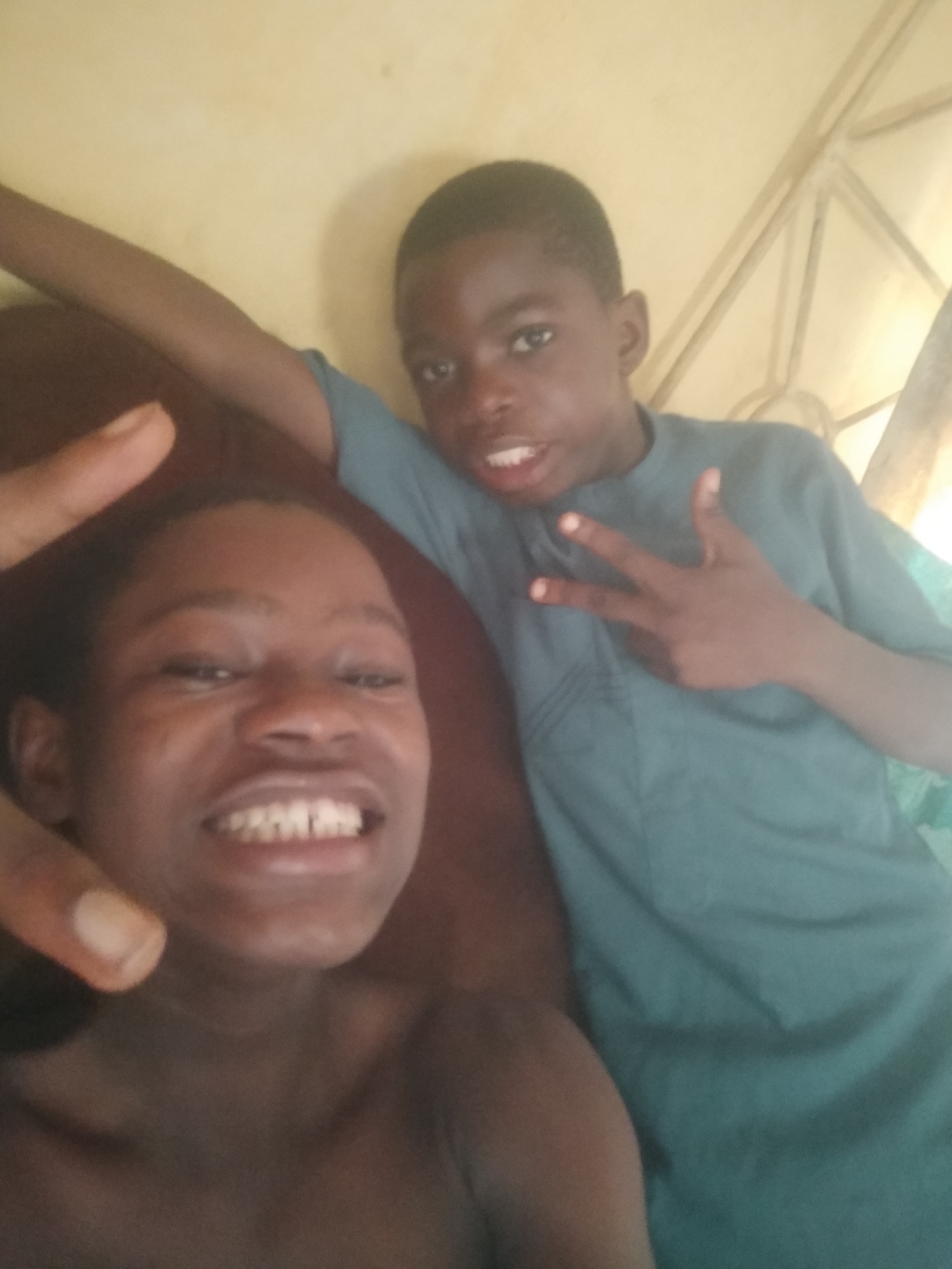 This close-up, casual photograph captures two young individuals, likely boys, in an indoor setting in Shangisha, Nigeria, during what appears to be daytime.
In the foreground on the left, a person with a dark skin tone is positioned very close to the camera, smiling broadly and revealing their teeth. Their expression conveys happiness and comfort. A finger, possibly a thumb, is partially visible in the upper left quadrant of the frame. This individual appears to be either shirtless or wearing a very dark garment that blends into the shadows.
Behind and to the right, the second person, also with a dark skin tone, is visible from the chest up. They are wearing a simple, short-sleeved teal or light blue shirt. This individual is making a peace sign with their left hand, directed towards the camera, and has their mouth slightly open. Their gaze is also towards the lens.
The background consists primarily of a light yellow or beige wall. Behind the person on the right, some faint, light-colored structural elements, possibly part of a window frame or bed frame, can be discerned. The overall impression is that of a friendly, relaxed moment, possibly a selfie or a casual photo taken in a home environment. There is no visible text in the image.