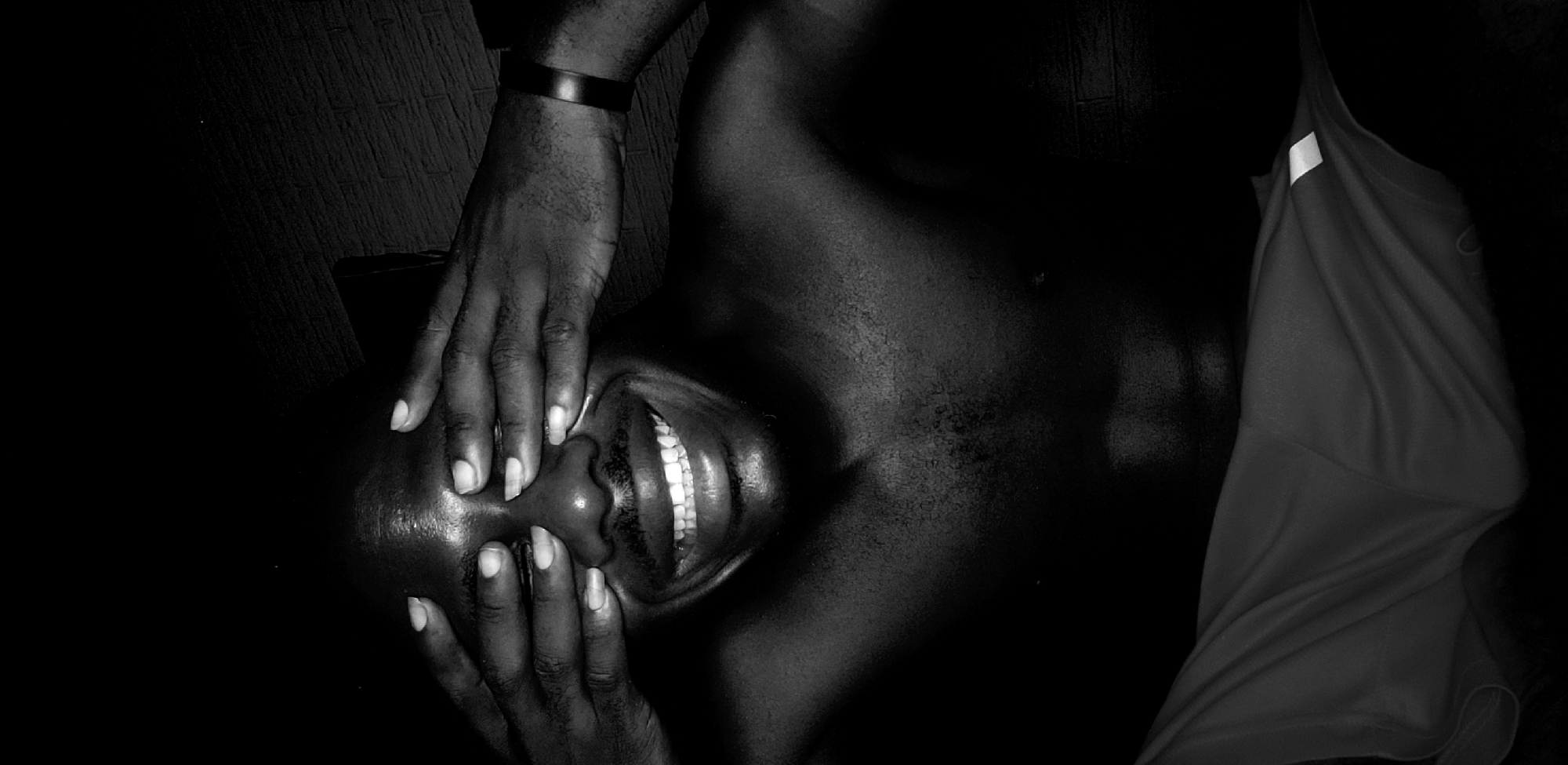 A high-contrast black and white photograph captures an intimate, horizontally-oriented close-up of a person with dark skin. The subject is smiling broadly, revealing a full set of bright white teeth, conveying joy or amusement. Two hands, with light-colored fingernails, are gently placed against the person's face, specifically cupping the cheeks and temples. A dark, band-like bracelet is visible on one wrist.
The image focuses on the person's head and upper torso, where the skin exhibits a distinct sheen, highlighted by dramatic lighting that creates strong contrasts and deep shadows. The background is largely obscured by darkness, with faint textures of a dark, possibly wooden, surface visible in the upper corners. On the right side of the frame, a piece of light-colored fabric, resembling a tank top or athletic wear, is draped, showing folds and a small, reflective stripe near its strap.
The overall impression is one of happiness and an intimate moment, amplified by the chiaroscuro lighting. No text is visible in the image. The location context provided is Narayi, Nigeria.