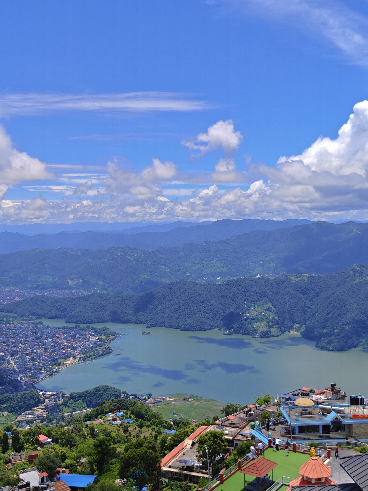 The image is a high-angle, wide shot capturing a panoramic view of Pokhara, Nepal, featuring the iconic Phewa Lake and surrounding mountainous terrain under a bright, cloudy blue sky. In the foreground, several buildings with flat roofs, some featuring rooftop seating areas and umbrellas, are visible. These structures appear to be hotels or restaurants, with people milling about on the rooftops, some seated at tables and others walking. The buildings themselves are a mix of white and blue, with some red-tiled roofs.
The mid-ground showcases the vast, placid Phewa Lake, its greenish-blue water reflecting the sky. A small island with a pagoda-like structure is situated in the lake. Along the western edge of the lake, the dense urban sprawl of Pokhara city is visible, characterized by a multitude of closely packed buildings with greyish rooftops. A few boats are dotted on the lake near the shore.
The background is dominated by lush, green, rolling hills and distant, hazy blue mountains, indicative of the Himalayan foothills. The landscape is covered in dense vegetation, with some terraced cultivation or scattered settlements visible on the hillsides. The sky is a vibrant blue with scattered cumulus clouds, some appearing wispy and others more voluminous, casting subtle shadows on the mountains. The overall impression is a picturesque, scenic vista of a city nestled amidst natural beauty.