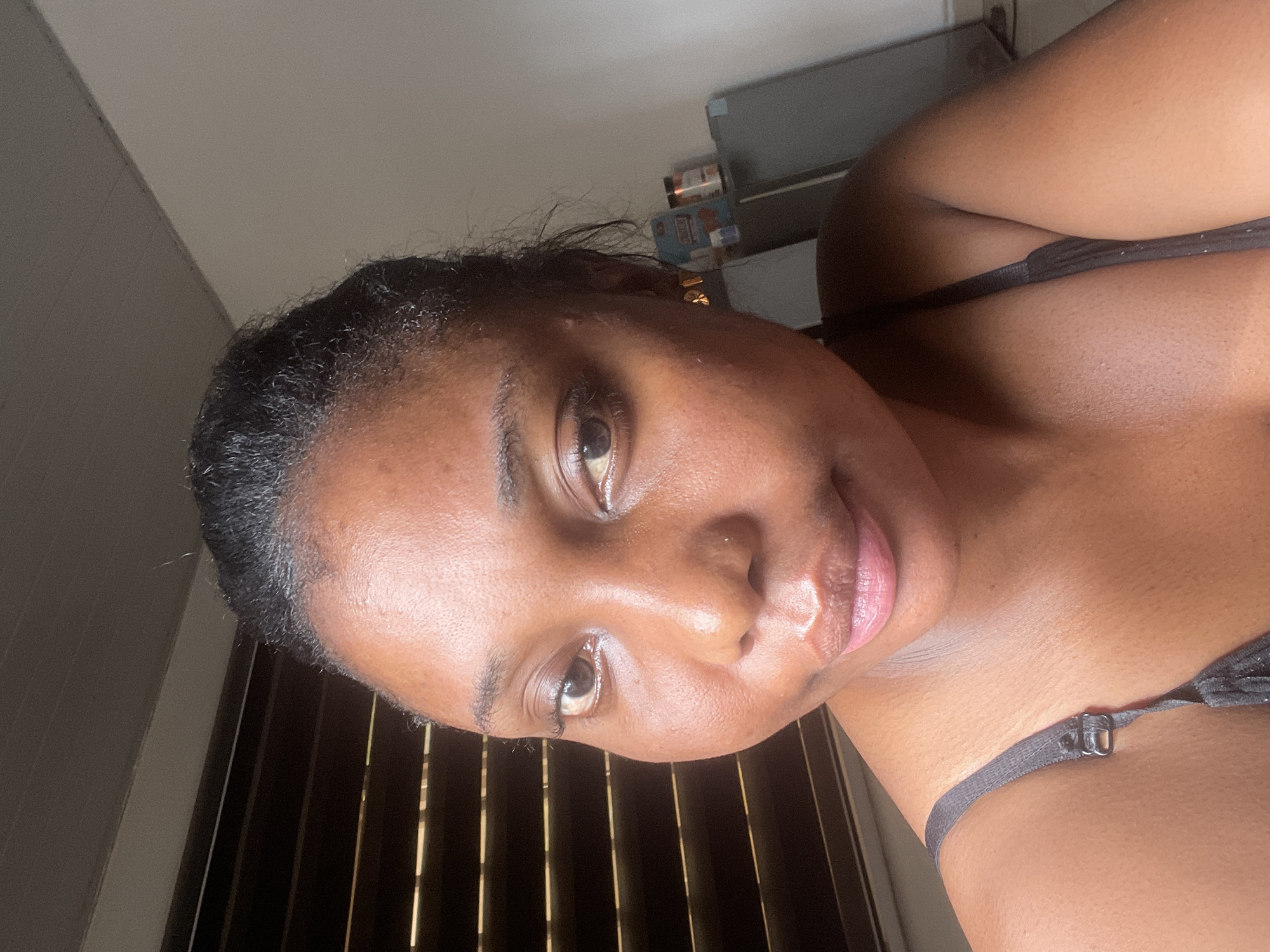 The image is a close-up selfie of a Black woman with short, dark hair. She is wearing a dark-colored top. The background includes a window with closed blinds and some objects on a shelf. The scene appears to be indoors. There is no information to suggest the location is in Abuja, Nigeria.