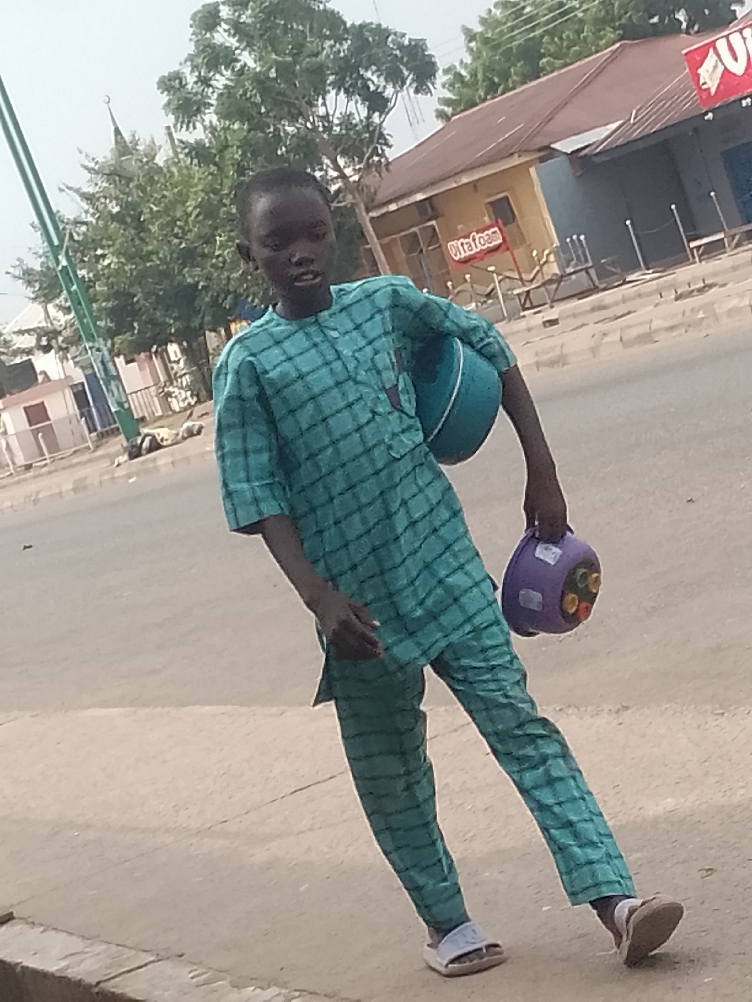 The image depicts a young boy walking on a sidewalk in Pantami, Nigeria. He is wearing a teal patterned traditional outfit and carrying a light blue bowl in his left arm and a purple container with colorful inserts in his right hand. The background shows a street scene with buildings, trees, and a traffic light pole. The boy is the central subject of the image, and his movement suggests he is walking somewhere. The environment appears to be an urban or semi-urban area with typical street infrastructure.