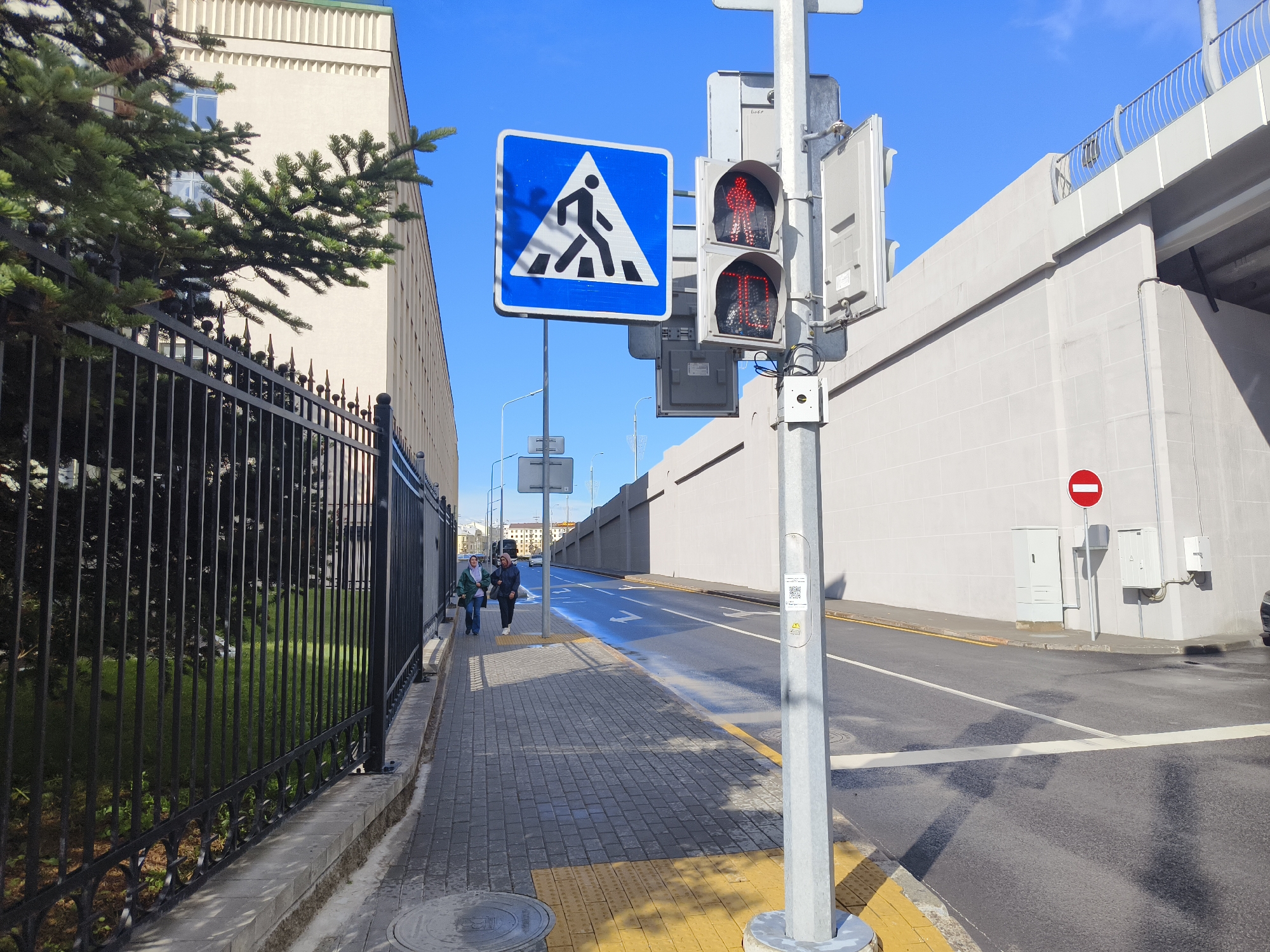 This media file captures a street scene in Minsk, Belarus. The primary subjects are a pedestrian crossing, a traffic signal, and a building. The traffic signal displays a red pedestrian icon, indicating that pedestrians should not cross. Adjacent to the signal is a blue and white pedestrian crossing sign.
The setting is an urban environment with a paved sidewalk and a road. A black metal fence runs along the left side of the image, separating the sidewalk from a grassy area and a tall, light-colored building. The building features multiple windows and a decorative textured facade. A mature evergreen tree is partially visible in the foreground.
In the background, a large concrete structure, possibly an overpass or bridge, dominates the upper right portion of the frame. A red circular sign with a white horizontal bar is visible on a light-colored wall to the right of the traffic signal, which is a "No Entry" sign.
Two people are walking on the sidewalk, moving away from the camera. They appear to be casually dressed, one wearing a green jacket and the other a darker jacket. The lighting suggests it is daytime, with the sun casting shadows. The sky is clear and blue, indicating fair weather. The scene appears calm and ordinary, with no specific event or heightened emotions evident.
Visible text includes a QR code and a warning symbol on the traffic signal pole, as well as potentially some text on the building in the background that is too small to read. The overall impression is a typical urban streetscape on a sunny day.