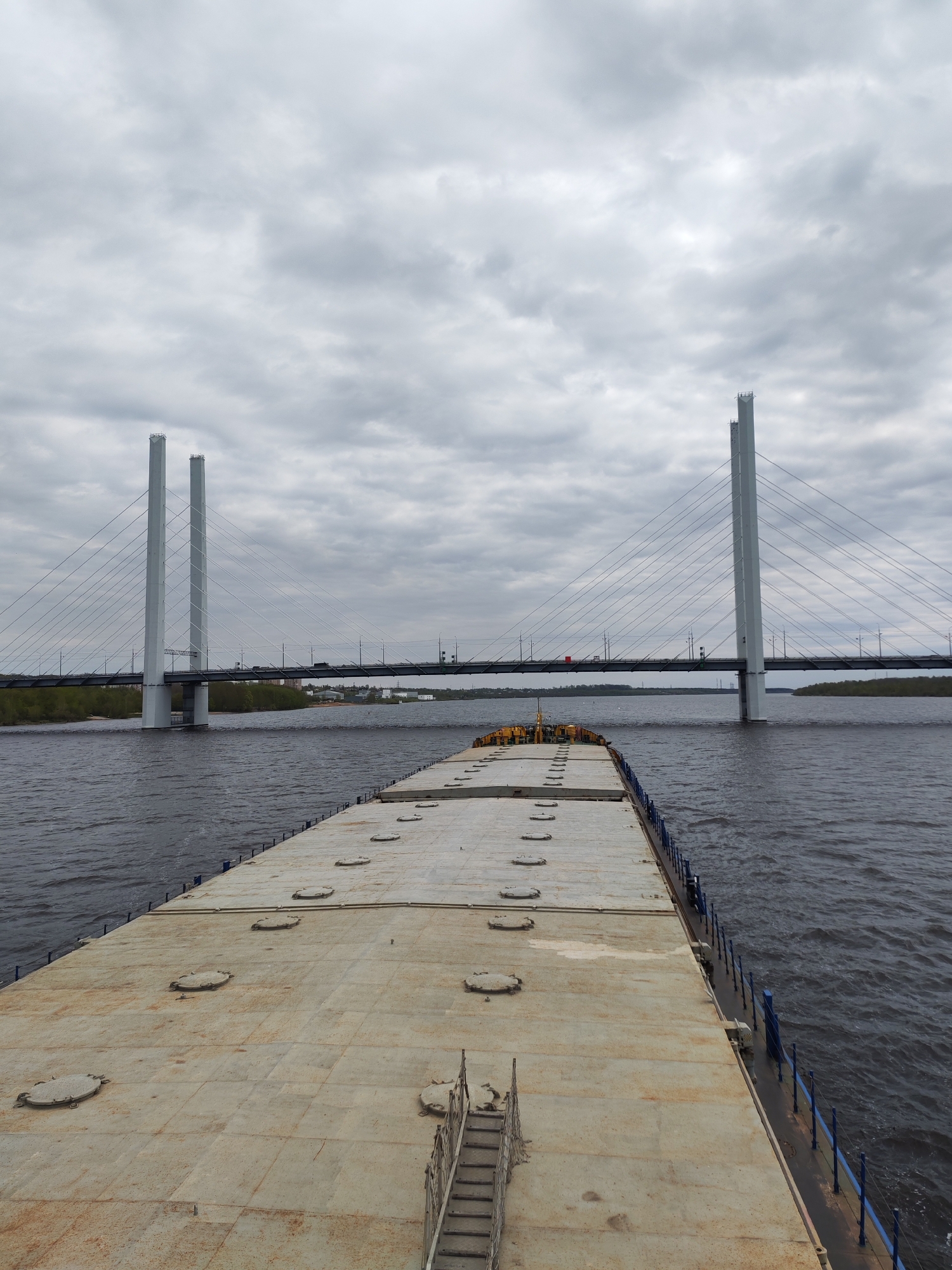 The image depicts a view from the deck of a barge, facing towards a cable-stayed bridge. The barge deck is flat, metallic, and has several circular access hatches. The bridge spans a wide body of water. The bridge towers are tall and white, supporting the bridge deck with numerous cables. On the bridge, some vehicles are visible. The sky is overcast with a dense layer of gray clouds. On the shoreline, vegetation and some buildings can be seen. The water is dark and choppy. The location of the city is unknown.