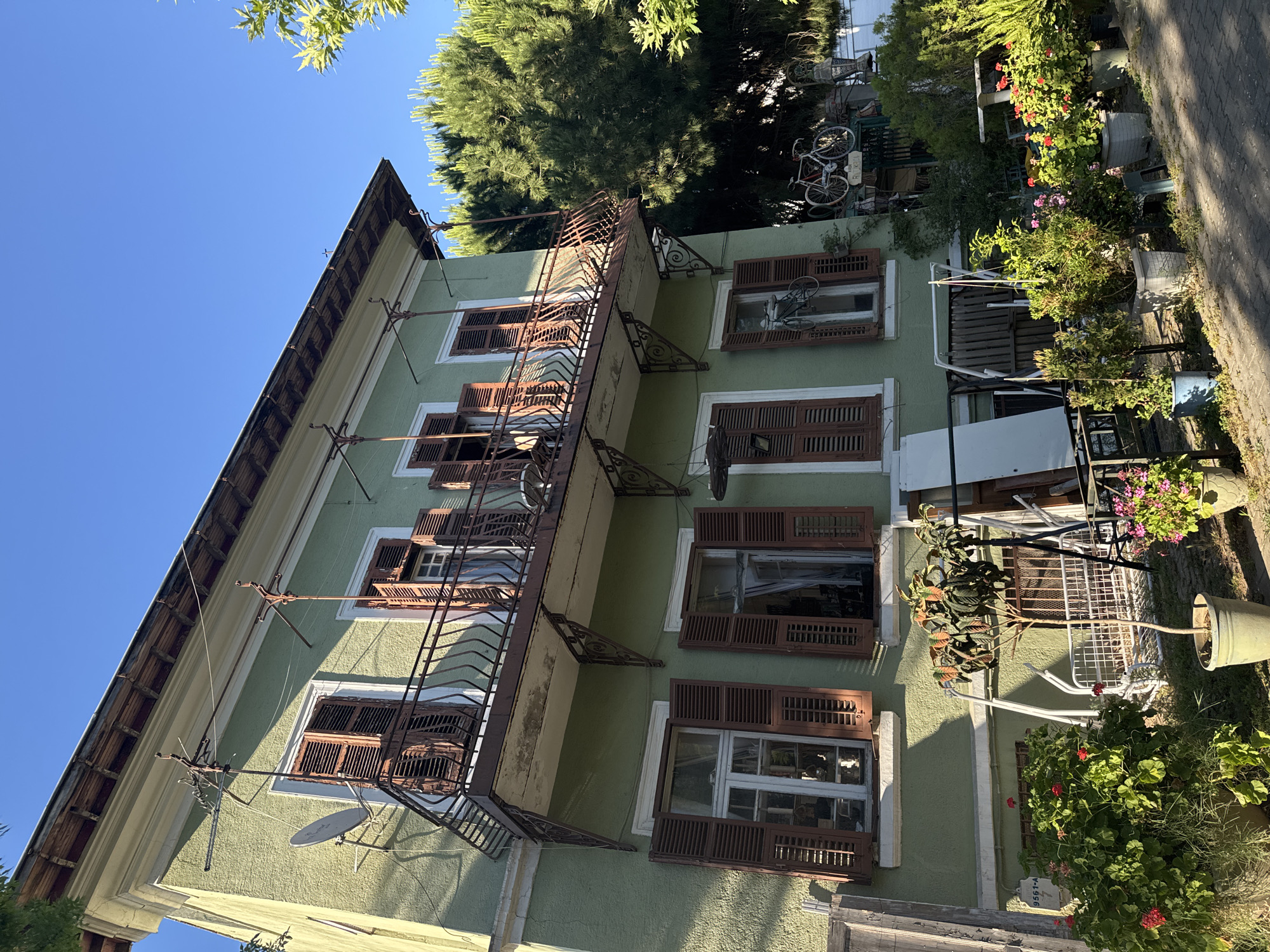 The image shows the exterior of a multi-story building, likely residential, painted in a light green hue. The building has several windows, many of which are fitted with dark brown wooden shutters. Some of the shutters are open, revealing the glass panes of the windows, while others are closed. A rusty, wrought-iron balcony or fire escape runs along the side of the building, with decorative metal brackets supporting it.
The scene is outdoors, with lush green trees and foliage visible around the building. The sky is a clear, bright blue, suggesting a sunny day. The lighting indicates it is daytime, with shadows cast by the building and surrounding objects. In the foreground, there are various potted plants, some with red flowers, and what appear to be outdoor furniture and possibly some discarded items, suggesting a lived-in, perhaps slightly cluttered, outdoor space. There is a satellite dish attached to the side of the building. In the background, on a higher level of the building or a nearby structure, two bicycles are visible.
The overall impression is that of a charming, somewhat weathered building in a residential area, likely in a warm climate given the abundant greenery and open windows. The location context provided places this scene on the Prince Islands in Turkey. There is some faint text on a white object in the lower right portion of the image, but it is too blurry to be legible.