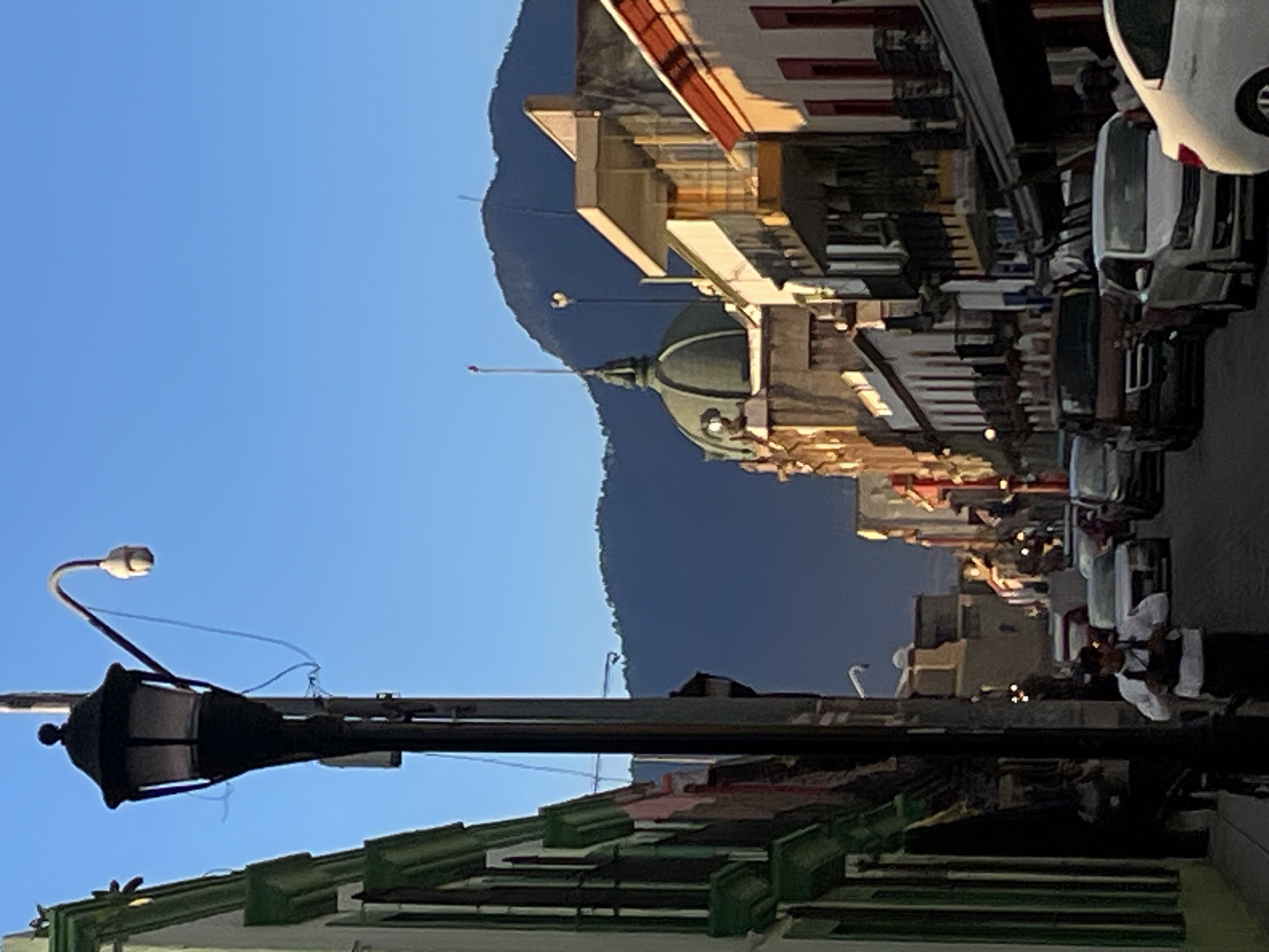 The image depicts a street scene in Oaxaca City, Mexico. In the foreground, a black, ornate street lamp stands out against a bright blue sky. A green-roofed building is partially visible beneath the lamp. In the midground, a domed building is situated in front of a large mountain. Further down the street, people are visible on the sidewalk. Parked cars line the right side of the street. The buildings lining the street appear to have colonial architecture with various colors. The light suggests it is either morning or late afternoon.