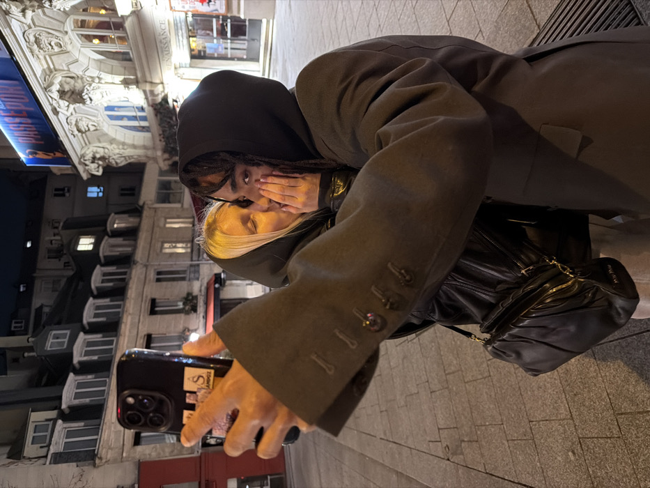 Two people are embraced closely on a city street at night. The individual in the foreground wears a dark hooded sweatshirt and has their hand over their mouth, a few dreadlocks visible. The other person, with blonde hair, appears to be kissing them on the forehead. The image is taken with a smartphone, held by an unseen person. Buildings with illuminated windows, some with ornate architectural details, are visible in the background, along with a glimpse of a theater sign.
