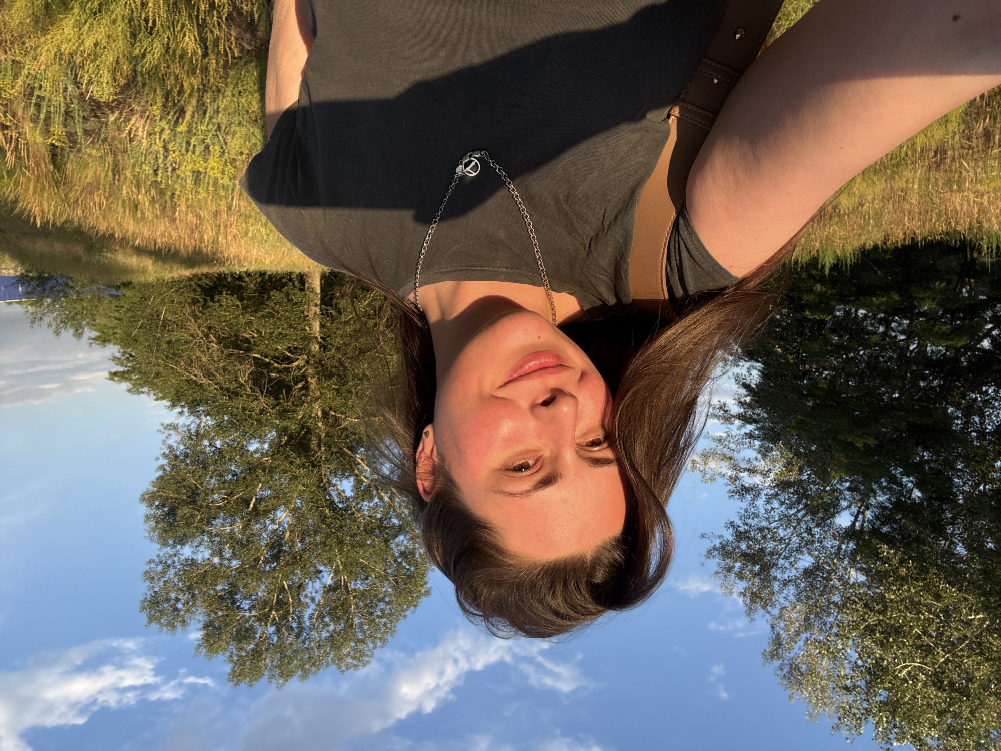 A woman is taking a selfie outdoors, with a bright blue sky and scattered clouds behind her. She has long, dark brown hair that is slightly windswept, and she is smiling softly at the camera. She is wearing a dark gray t-shirt and a silver necklace with a circular pendant. A brown bag strap is visible over her left shoulder. The background consists of a field of tall, dry grass and yellow wildflowers, with several trees in the distance. The lighting suggests it is late afternoon or early evening, with the sun casting a warm glow on the scene. There are no specific landmarks or identifiable structures that would suggest a particular city or location within a city.