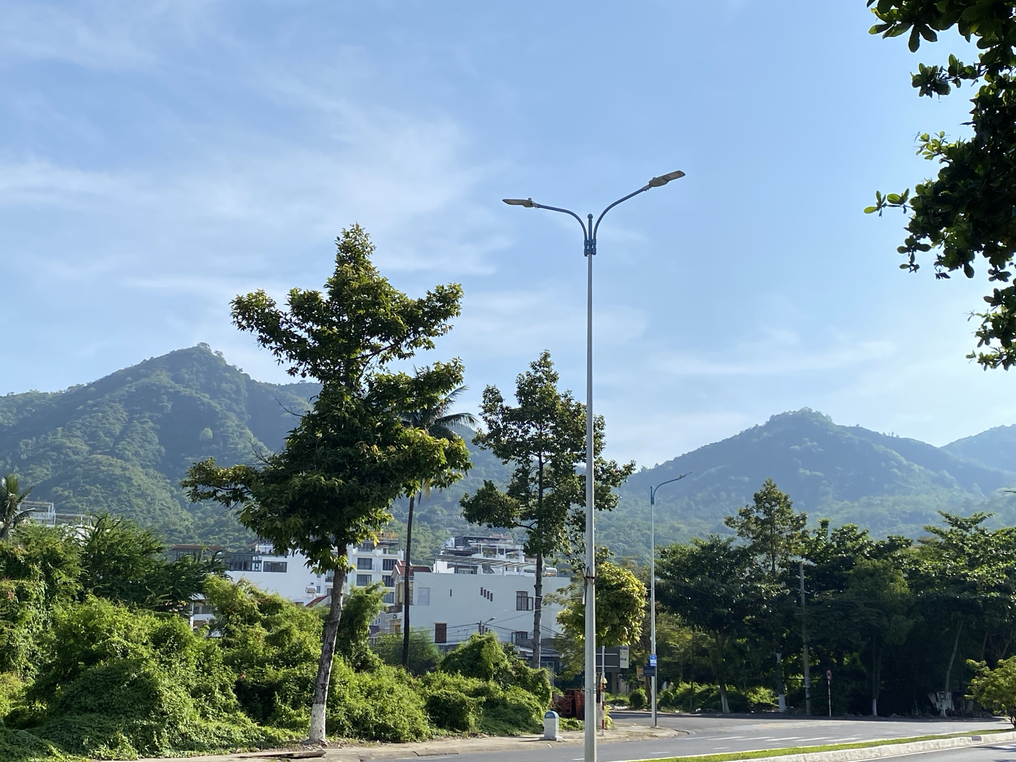 The image depicts a street scene in Bac Nha Trang Ward, Vietnam, characterized by lush greenery and mountainous terrain. In the foreground, a paved road with white lane markings runs horizontally across the frame. A single grey streetlamp with two lights stands prominently in the center-right of the image. To the left of the streetlamp, several trees with dense green foliage are visible. Behind these trees, a multi-story white building with dark-framed windows is partially obscured by vegetation. In the background, rolling green hills and mountains dominate the landscape under a clear blue sky with scattered clouds. The overall atmosphere is serene and natural, with a touch of urban development. There are no people or animals visible in the image.