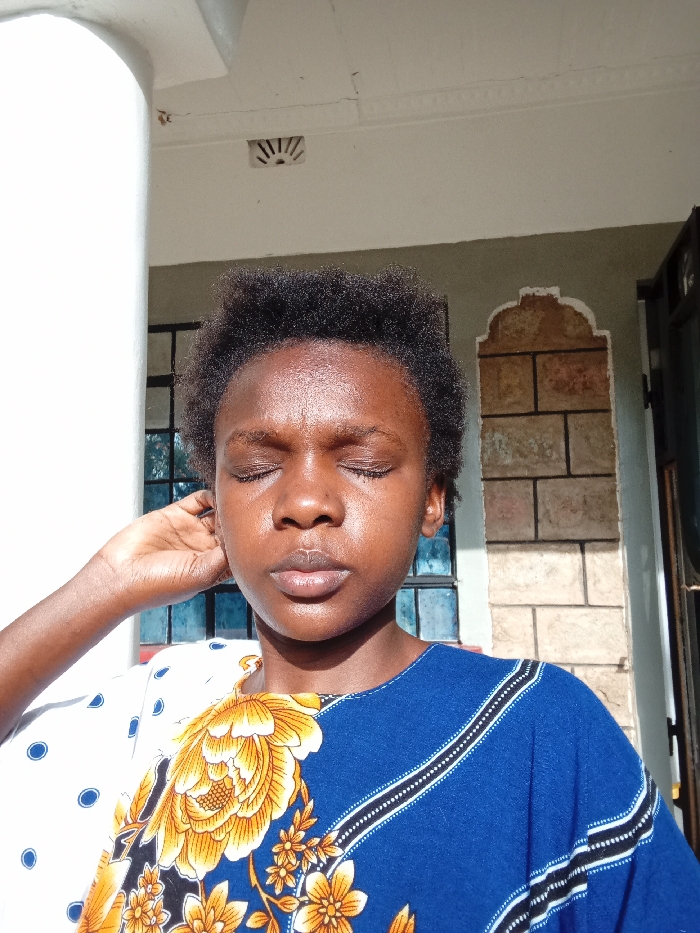 An individual with dark skin and short, dark, curly hair is depicted in a brightly lit outdoor or semi-outdoor setting within Mahiakalo ward, Kenya. Their eyes are closed, and their face exhibits a sheen consistent with direct sunlight exposure. The individual's right hand is raised, with fingers gently curled, positioned near the right ear/temple area. They are wearing a garment featuring multiple patterns: a white section with blue polka dots on the viewer's right shoulder, a central section with large yellow and orange floral designs, and a dark blue section with diagonal white and black striped patterns on the viewer's left shoulder.
The background includes a thick, white, rounded column on the far left. Above, a light-colored ceiling or overhang shows minor cracking and a small, semicircular vent with radiating slats. A light grey-green wall is visible, featuring a window with dark frames and blue-tinted panes to the viewer's left of the individual. To the viewer's right, an arched architectural feature with visible, rectangular, light brown stone blocks is present on the wall. A dark-framed door or window is partially visible on the far right. Strong, direct sunlight illuminates the scene from the viewer's right.
