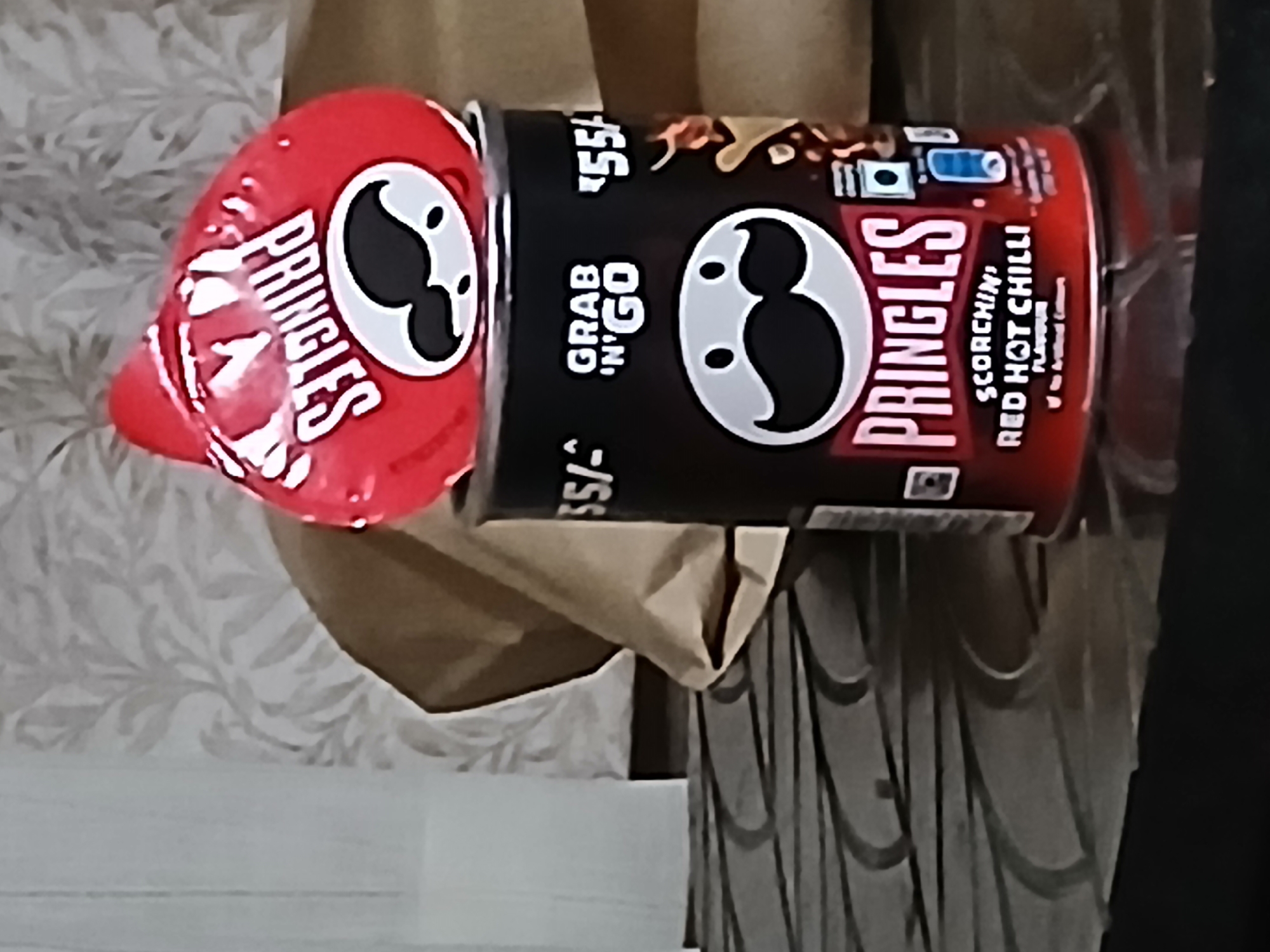 A red and black Pringles can is prominently displayed, specifically labeled "Scorchin' Red Hot Chilli". Next to it is a smaller, red Pringles container. The scene appears to be indoors, as evidenced by the wall and the bag underneath the Pringles. The setting suggests the presence of Pringles, which are the focus of the image. There is no indication of Bengaluru or any people in the shot.