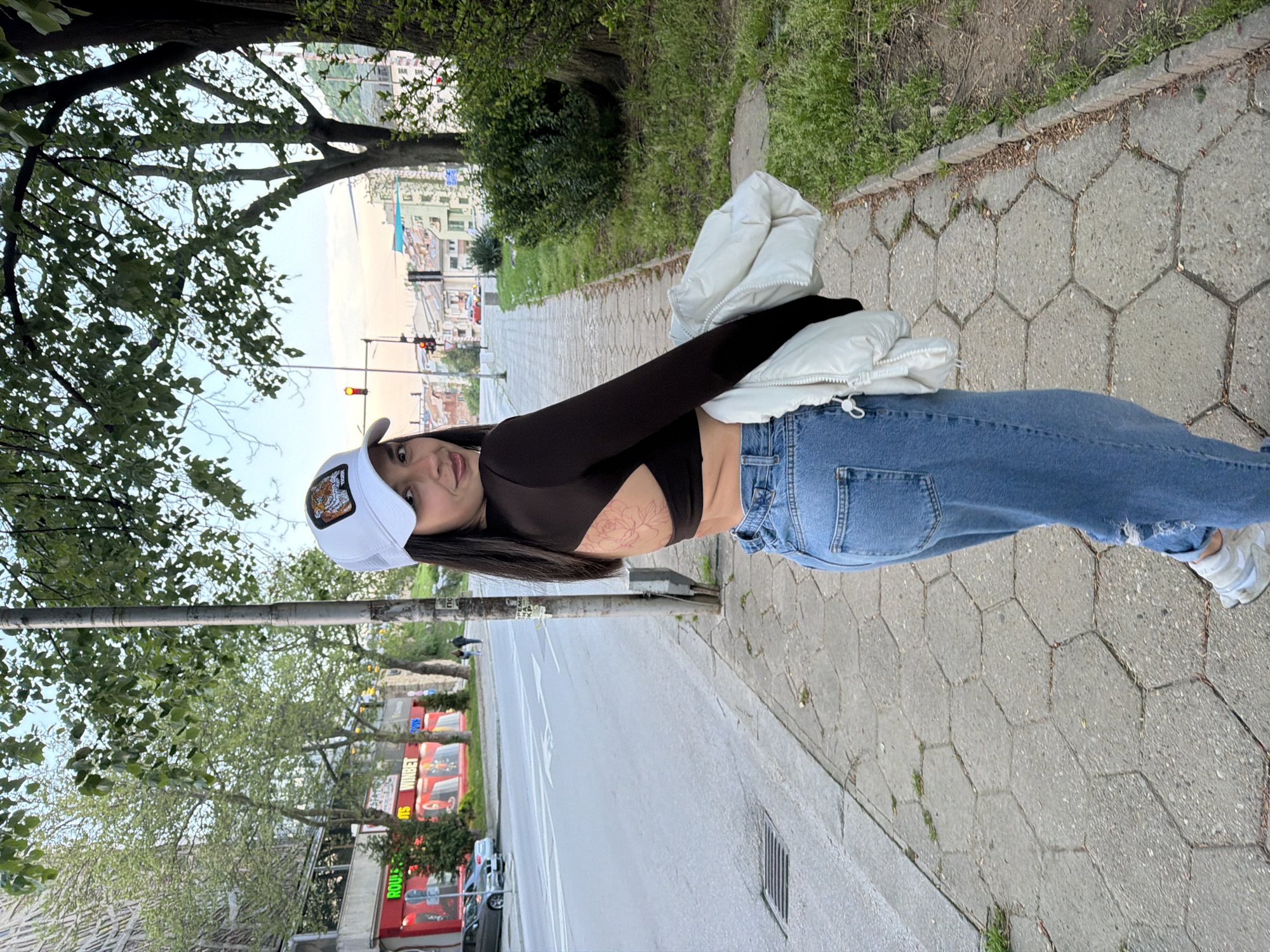 A young woman stands on a sidewalk in what appears to be an urban environment. She is wearing a black long-sleeve top that exposes her midriff, blue jeans, and white sneakers. She also wears a white baseball cap. A white jacket is draped over her arm. A tattoo is visible on her side.
The sidewalk is paved with hexagonal concrete tiles. Behind her is a street with trees lining the sidewalk, and beyond the street, there are buildings and traffic lights. One of the buildings has the brand "Roulette" displayed prominently. The buildings and street details suggest this location is Gabrovo, Bulgaria.