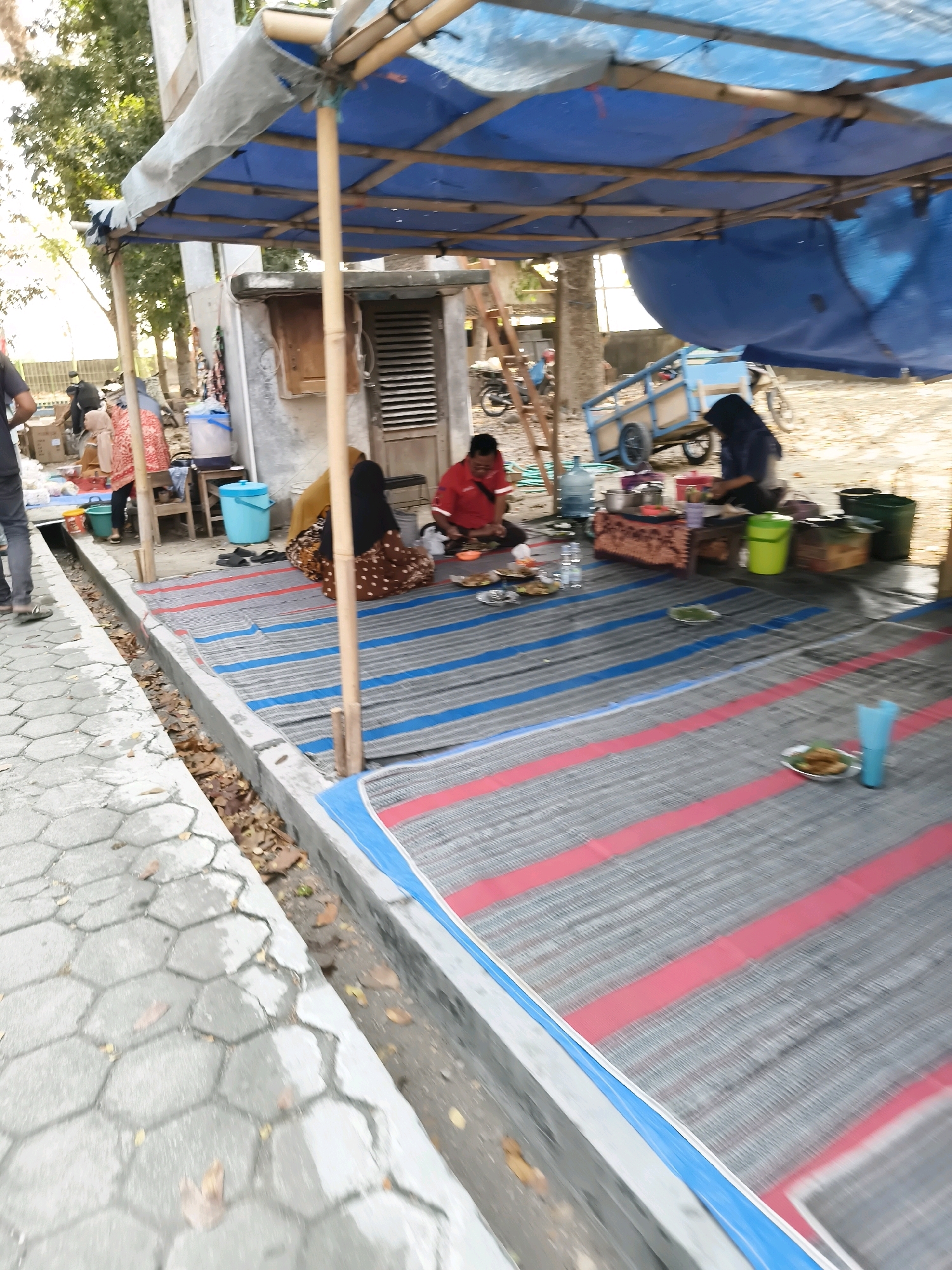 This is a ground-level, medium-shot, outdoor photograph taken in Tlogorejo, Indonesia, depicting a scene under a makeshift canopy constructed from blue tarp and bamboo poles. The canopy covers an area where several people appear to be eating or socializing on woven mats laid on the ground. In the foreground, a grey, hexagonal paved walkway runs alongside a shallow drainage ditch containing fallen leaves and debris. Behind the walkway, within the shaded area, two individuals are seated on a mat. One person, wearing a red shirt, is facing away from the camera and appears to be eating from a plate. Another individual, wearing a dark headscarf and patterned clothing, is seated with their back to the camera, also engaged in an activity at a low table. Other individuals and various objects are visible in the background, including a blue water cooler, buckets, and a small cart. The overall impression is of an informal gathering or a temporary market setup.