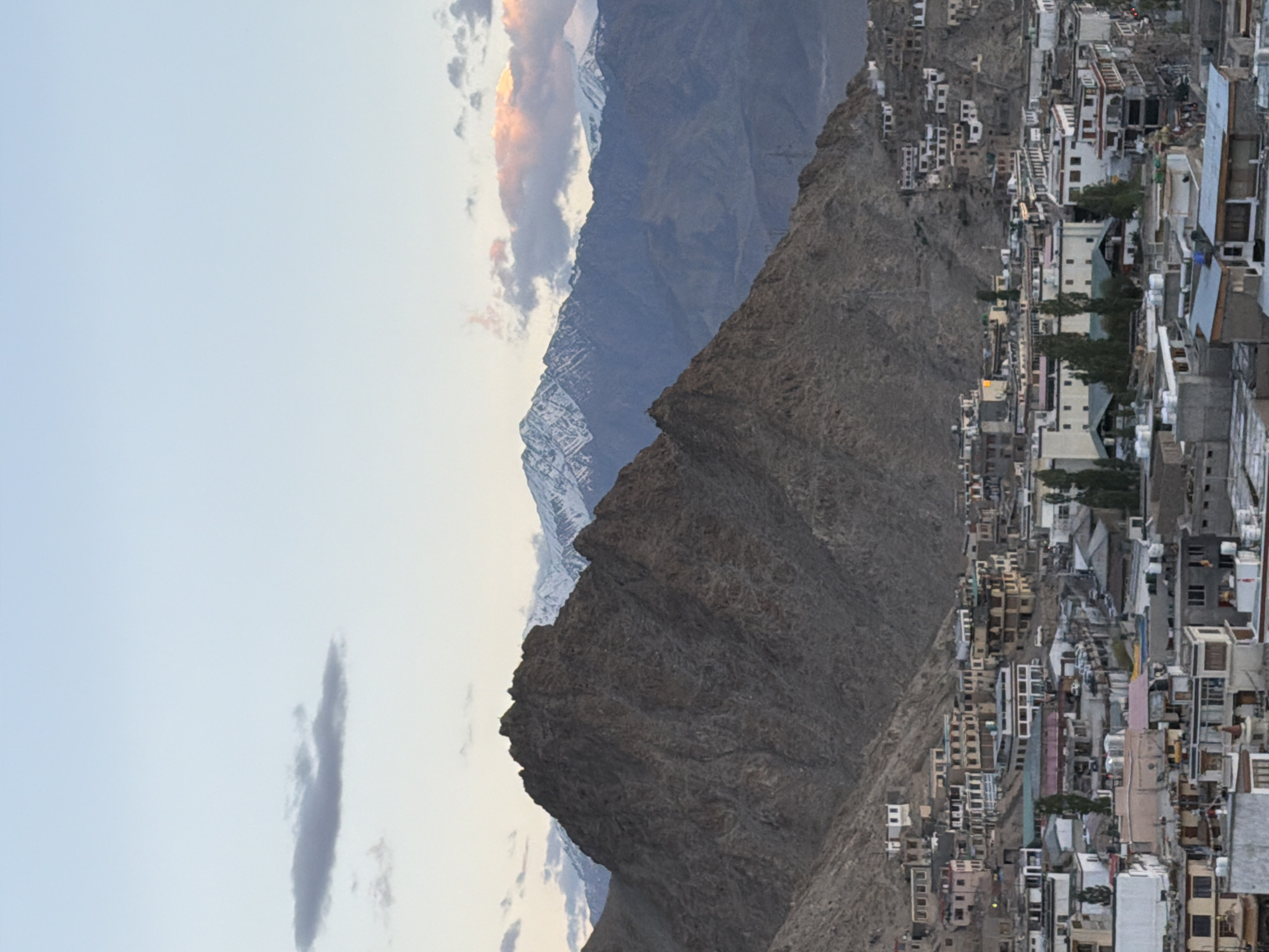 The image depicts a view of the city of Leh, India, nestled against a dramatic mountainous landscape. In the foreground, a dense collection of buildings, predominantly in muted tones of white, beige, and brown, are visible. The architecture appears to be a mix of traditional and modern styles, with flat roofs and multiple stories. Some buildings are clustered together on hillsides, indicating the city's elevation. Scattered trees are interspersed among the structures.
Behind the city, a massive, rugged, and rocky mountain dominates the mid-ground. Its steep slopes are devoid of vegetation, showcasing the arid terrain. Further in the background, a range of higher mountains with snow-capped peaks stretches across the horizon. The sky above is a gradient of soft blues and pale grays, with a few wispy clouds. One cloud on the left has a darker, more defined shape, while others higher up are tinged with the warm colors of sunrise or sunset. The overall impression is one of a high-altitude settlement in a remote, mountainous region. There are no discernible people or specific actions taking place in the image.