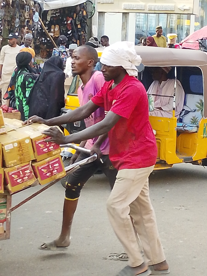 The scene depicts a street in Fagge A, Nigeria, during daylight. Two dark-skinned males are transporting goods on a metal handcart from left to right. The lead male wears a red short-sleeved shirt, light-colored trousers, and a white headwrap; his hands grip the cart's handles. The handcart carries multiple golden-brown cardboard boxes, each marked "GOLD" with red and white details, stacked in at least three visible tiers. The second male, positioned behind and to the left of the first, wears a pink short-sleeved shirt, dark shorts with yellow bands on his left leg, and sandals.
Immediately to the right of the handcart, a yellow three-wheeled auto-rickshaw contains at least two passengers, dressed in light-colored traditional garments. The background includes a bustling market environment. On the far left, a market stall displays numerous sandals. Further back, a light-colored building features "SUPER STORES" and "pure" signage. Multiple other individuals are visible throughout the scene, including a woman in a black hijab and patterned dress behind the cart, and various men in traditional and casual attire. The ground is paved.