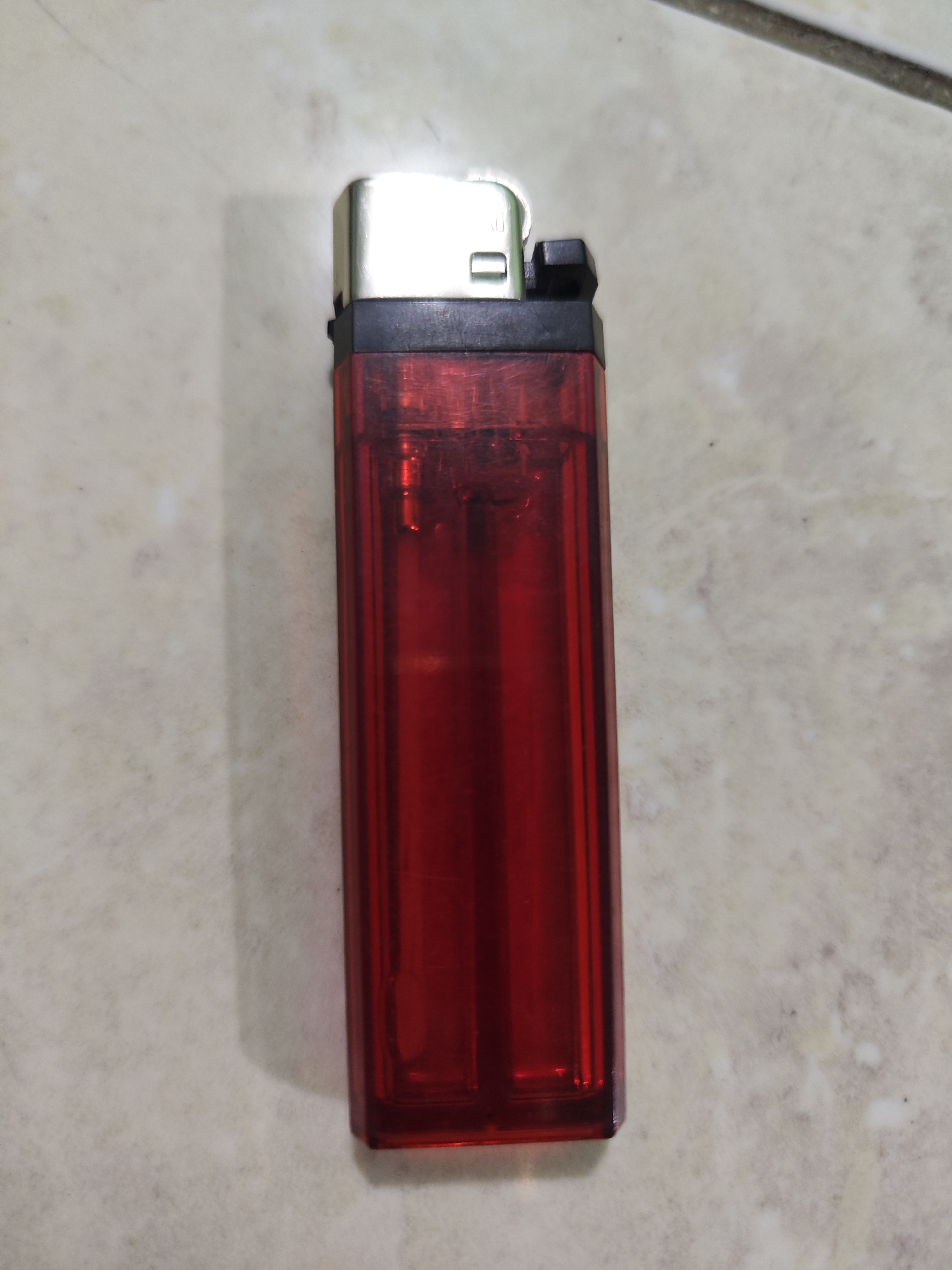 A red disposable lighter lies on a light-colored, textured surface, possibly a tiled floor. The lighter is oriented vertically, with the metallic cap at the top and the transparent red body below. The cap is silver-colored with a black plastic mechanism visible. The red body of the lighter appears to be filled with a liquid, likely butane, indicated by a slight sheen. The surface the lighter rests on has a subtle pattern, suggesting it is indoors or on a paved outdoor area. There is a shadow cast to the left of the lighter, indicating a light source from the right. The overall scene is still and static, with no people or actions depicted. The specific location within Sroyo, Indonesia is not visually inferable from the image.