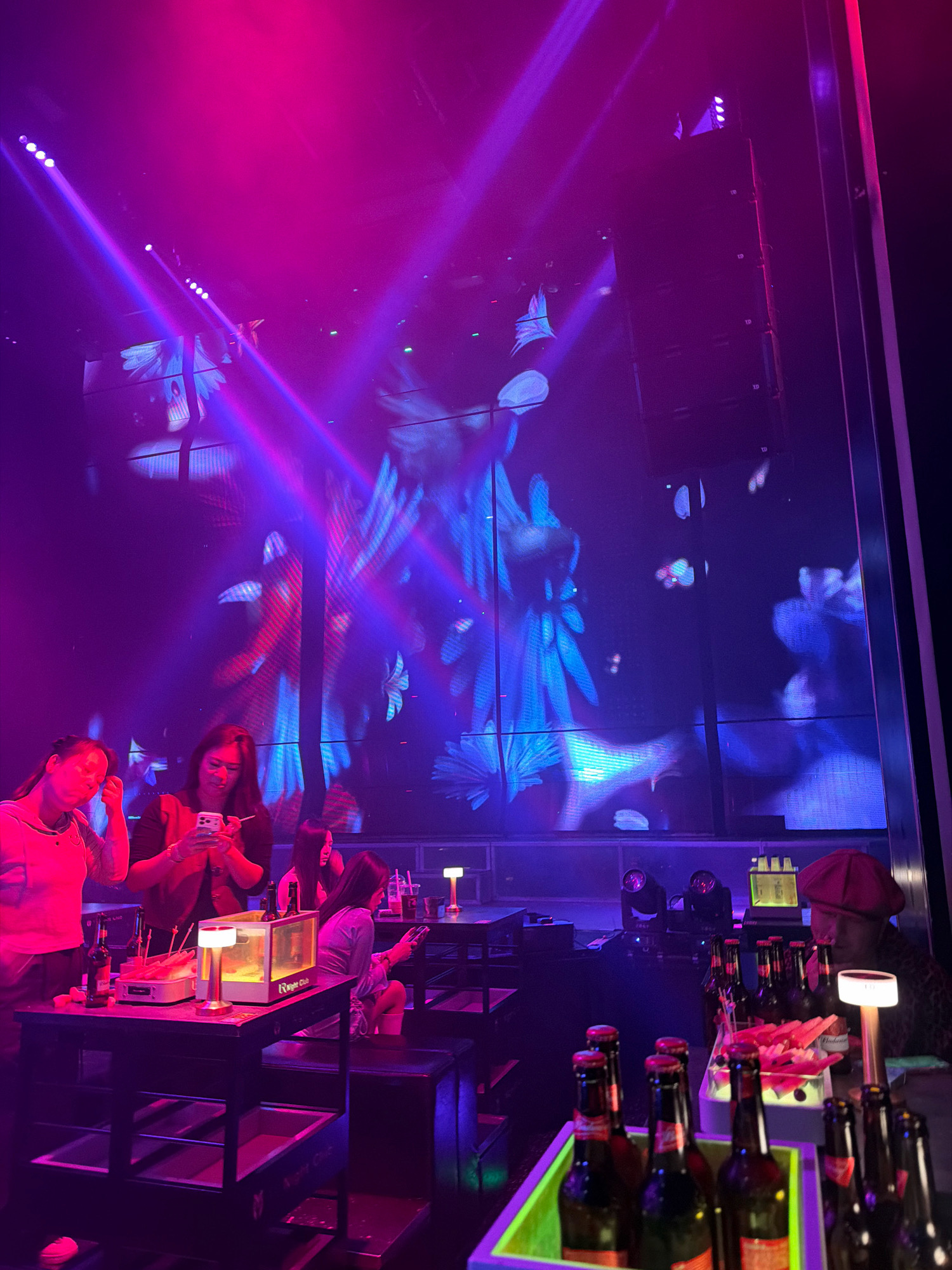 The image depicts a vibrant nightclub scene in Hunchun, China, illuminated by bright pink and purple lights and a large screen displaying abstract blue and red visuals. In the foreground, a group of women are gathered around tables, engaged with their phones. One woman in a pink top holds up her phone, while another in a dark jacket uses hers while standing. A woman in a light purple outfit is seated, also looking at her phone. On a table in the center, there are drinks, snacks, and a small lamp. In the background, more tables and patrons are visible, along with stage lighting and speakers. The atmosphere is lively and typical of a nighttime entertainment venue.