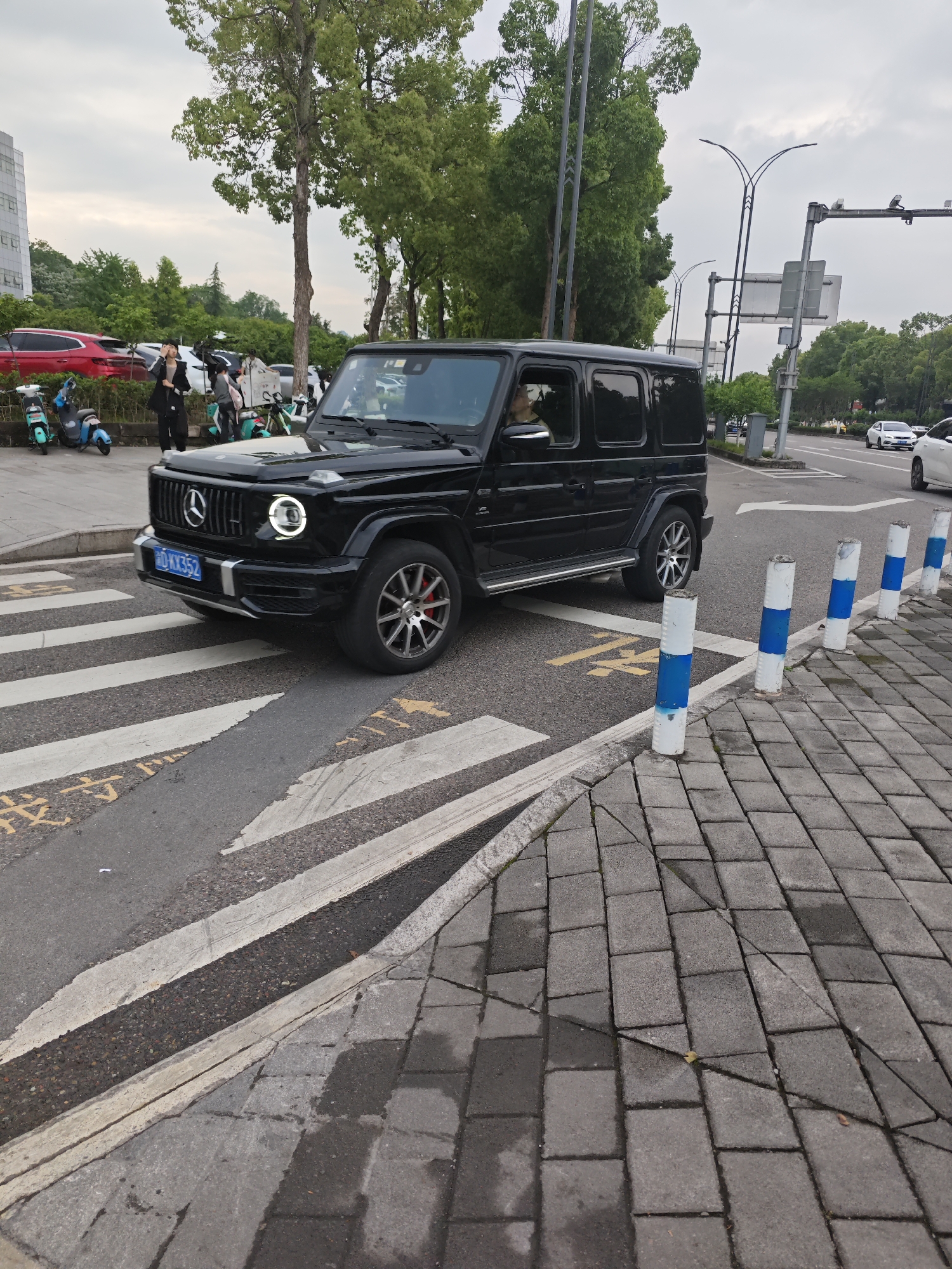 A black Mercedes-Benz G-Class SUV is positioned on a crosswalk in Chongqing High-tech Zone, China. The SUV has its front driver's side wheel on the white striped portion of the crosswalk, with the front end also on the crosswalk. A woman is standing on the sidewalk near a group of parked scooters and a red car behind the SUV. Street lights and trees are visible along the road. The curb is lined with white posts featuring blue bands. The road is asphalt and the sidewalk is paved with gray bricks. Another white vehicle is on the road further in the distance.