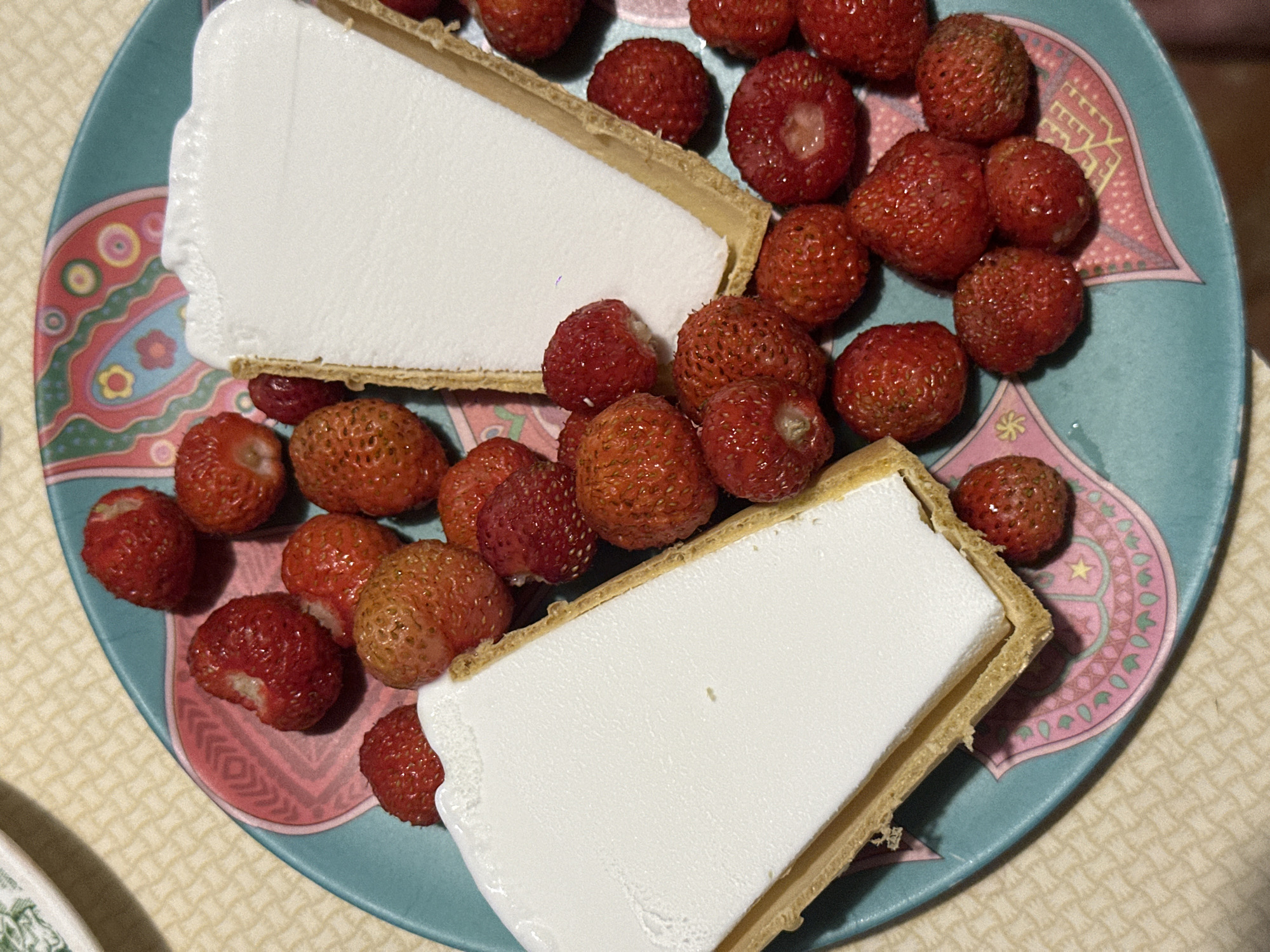 The image shows a close-up of a teal-colored plate filled with fresh strawberries and two slices of ice cream tarts. The tarts have a golden-brown crust and are filled with a white, creamy substance, likely ice cream or a similar frozen dessert. The strawberries are scattered around the tarts, some whole and some with small bites taken out of them, suggesting they are being eaten.
The plate has a decorative pattern with pink and light blue geometric shapes and floral motifs. The background appears to be a light-colored, textured tablecloth. The lighting suggests it might be daytime, possibly indoors, given the diffuse light and lack of harsh shadows. The overall scene conveys a sense of a casual dessert offering, possibly a snack or a light dessert after a meal. There are no people visible in the image, and no text is present. The presence of fresh strawberries and a sweet dessert hints at a pleasant, perhaps summery, occasion. Given the location context of Biysk, Russia, this could be enjoyed during the warmer months when strawberries are in season.