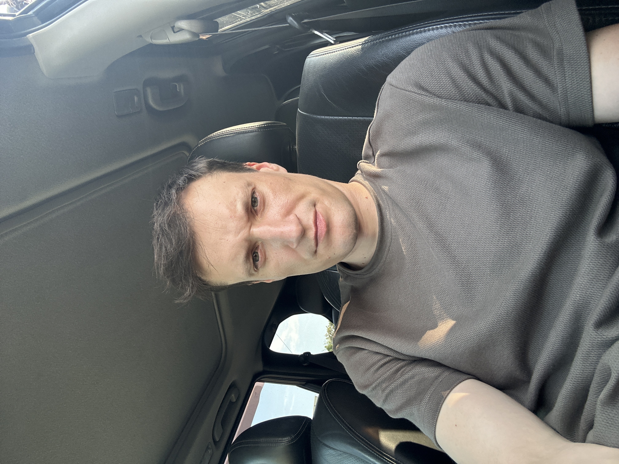 A man is seated in the driver's seat of a car, looking directly at the camera. He has short, dark hair and is wearing a brown, short-sleeved t-shirt. The interior of the car is visible, including black leather seats and the car's sunroof. Sunlight streams in from the windows, casting shadows on the man's face and shirt. The background outside the car windows shows foliage and a bright sky, suggesting an outdoor location. The image was taken in Shatava, Ukraine.