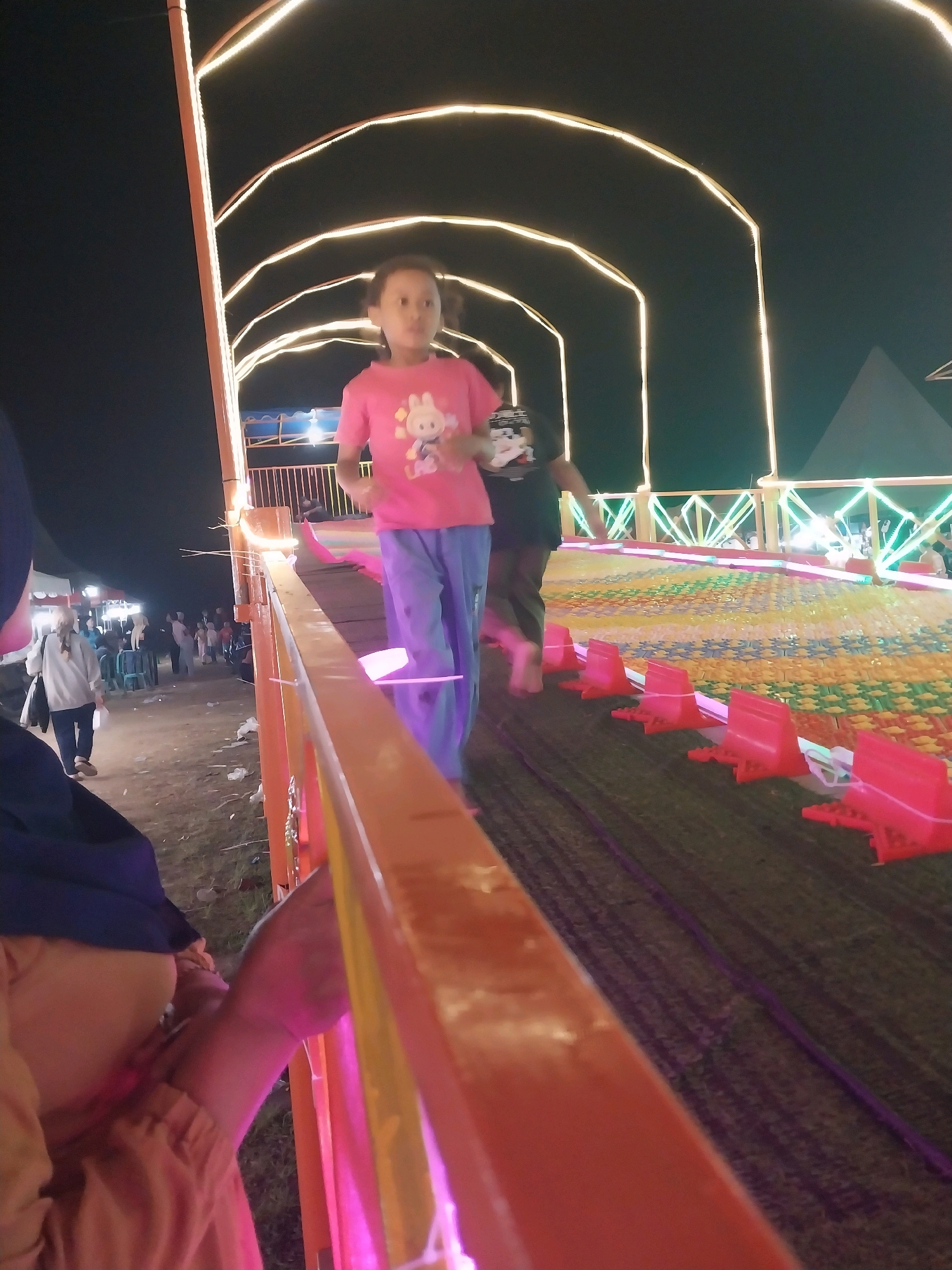 In the city of Jiwut, Indonesia, a nighttime scene unfolds at a fairground. A young girl in a pink t-shirt and blue pants is captured mid-stride on a colorful, patterned walkway. The walkway is lined with vibrant pink barriers. Overhead, a series of illuminated arches, outlined with yellow lights, form a tunnel-like structure, casting a warm glow. In the background, a crowd of people mill about, some walking and others standing near tents and stalls, suggesting a lively atmosphere. The ground appears to be a mix of dirt and scattered debris. The scene is illuminated by artificial lights, including the decorative arches and lights from the fairground attractions.
