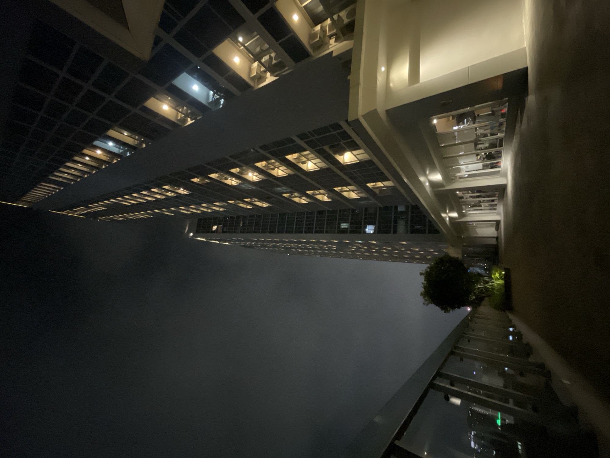 The image depicts a nighttime view looking up at a modern skyscraper in Central Jakarta, Indonesia. The building is illuminated, with many windows lit, suggesting it is occupied. The perspective is from a lower vantage point, looking upwards and slightly across the building's facade.
The scene is set at night, as indicated by the darkness and the artificial lighting of the building and the city below, which is reflected on the wet-looking ground in the foreground. The weather appears to be misty or foggy, obscuring the view of the lower parts of the city and creating a soft backdrop for the illuminated building.
In the foreground, there is a balcony or elevated walkway with a glass railing. Beyond the railing, a small, well-maintained tree is visible, as well as what appears to be another section of the building or an adjacent structure with more lights. The overall mood is one of urban sophistication and the quiet energy of a city at night. No people are clearly visible, and no text is present in the image.