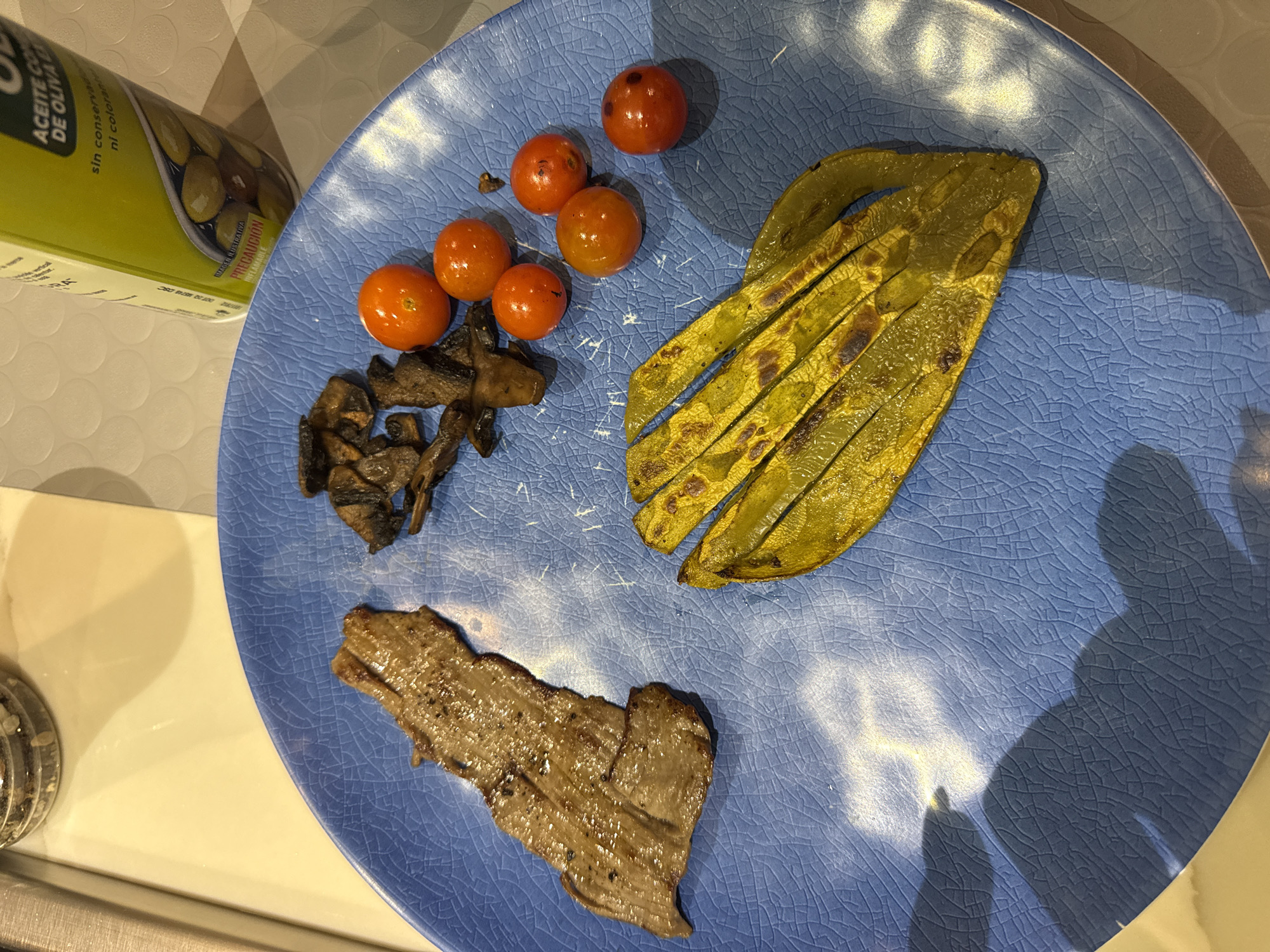 A blue ceramic plate holds a meal consisting of a portion of grilled meat, mushrooms, cherry tomatoes, and grilled nopal (cactus pad). The plate rests on a light-colored surface, near a can of olive oil. The location, San Bernardino Tlaxcalancingo, Mexico, is derived from text on the can of olive oil in the top left corner of the image.