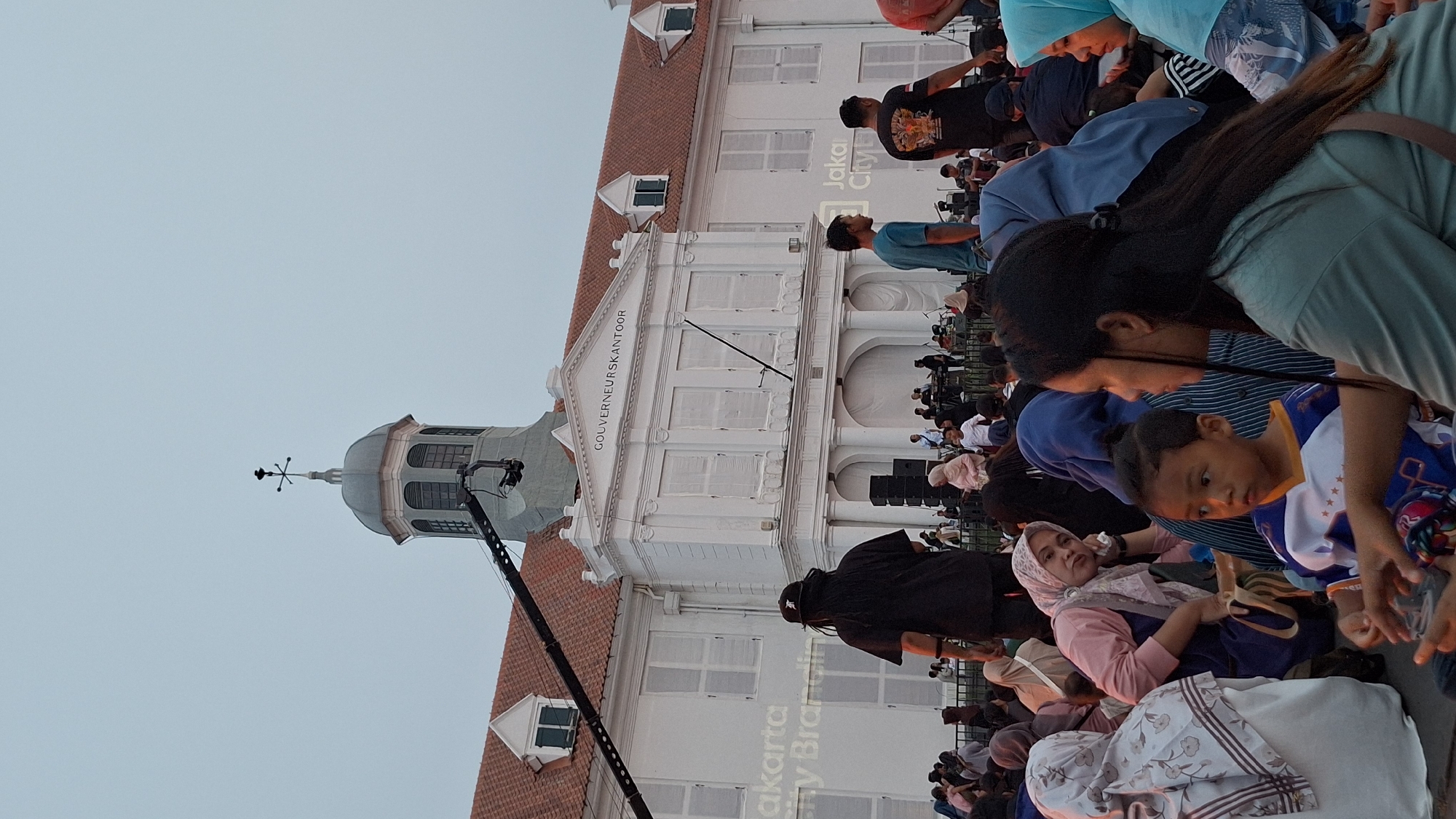 The image depicts a scene in front of the Gouverneurskantoor building, located in the Fatahillah Square of the Kota Tua area in West Jakarta, Indonesia. A large crowd of people is gathered outdoors, sitting on the ground, suggesting an event or performance is taking place. In the background, the white colonial-era building with a distinctive dome and clock tower stands prominently. A stage with musical equipment and performers is visible in front of the building. A large camera crane is positioned on the left side of the frame, indicating that the event is being filmed. The foreground features a group of people, including several women and a young boy, some of whom are wearing headscarves. The overall atmosphere appears to be relaxed and communal, with people enjoying the outdoor gathering.