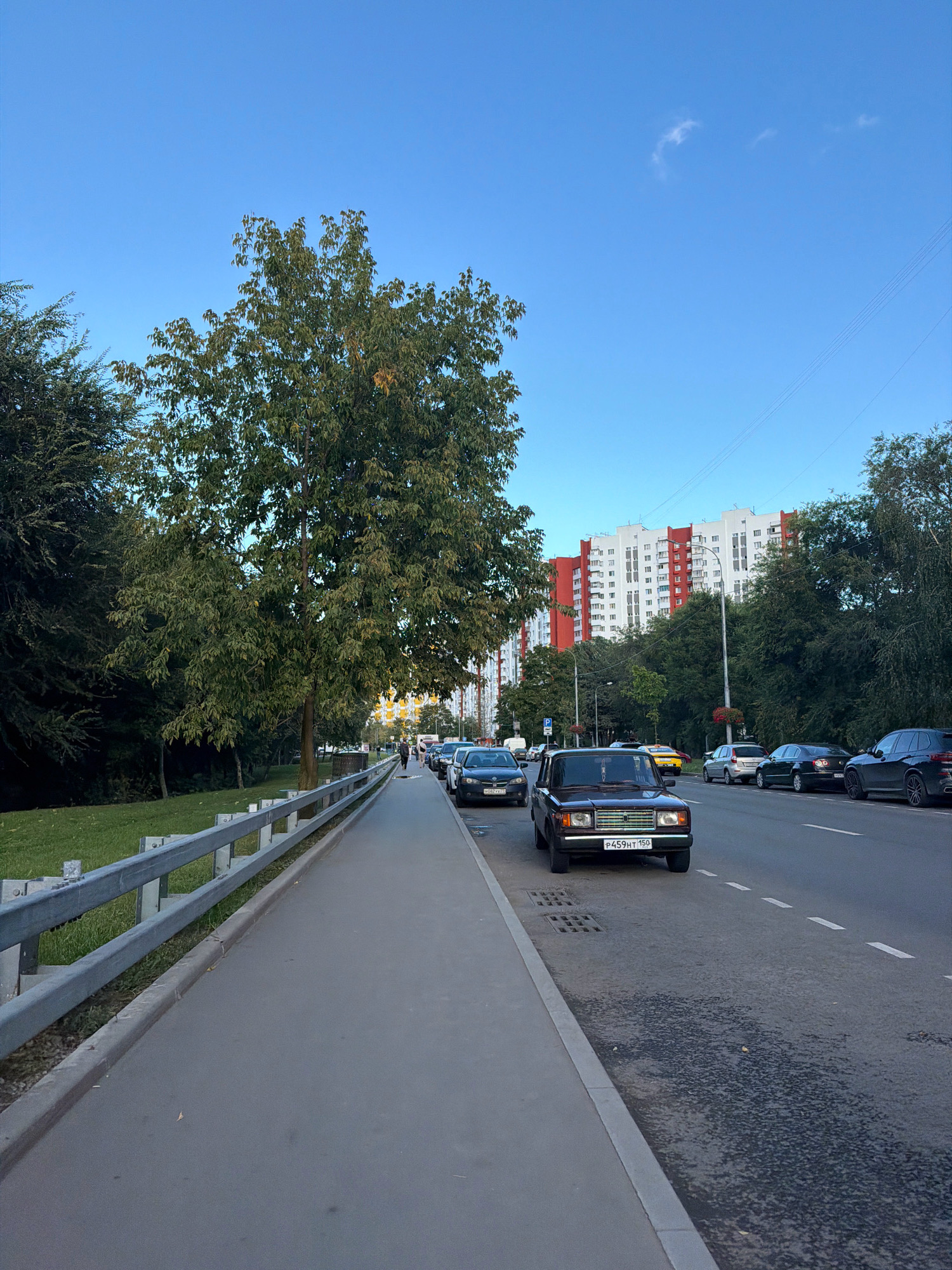 The image depicts a street scene in Moscow, Russia, on a clear day with a bright blue sky. A paved pedestrian walkway runs along the left side of the frame, separated from a grassy area by a metal guardrail. Several cars are parked along the right side of the street, and a steady stream of traffic moves in the same direction. In the background, a large residential building with white and red facades stands tall, surrounded by trees. The overall impression is one of an urban environment, possibly a residential neighborhood, with typical city infrastructure and activity.