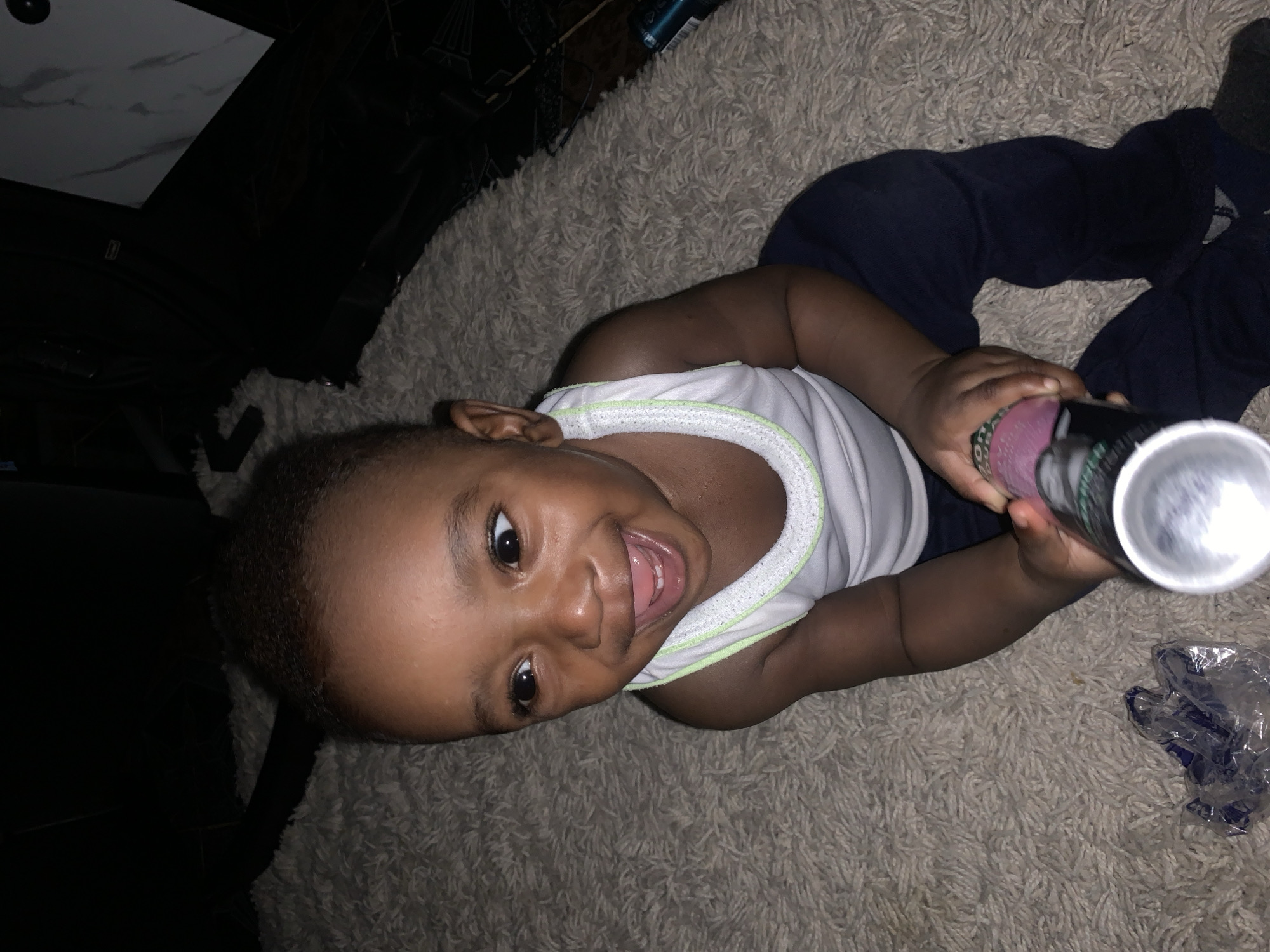A Black toddler sits on a cream-colored shag carpet. The toddler is wearing a white tank top with light green trim and navy blue pants. The toddler is holding a silver can with a pink and green label in both hands. The toddler's head is turned to the left, and they are smiling with their mouth open, showing teeth and tongue. Their eyes are wide and looking upwards. In the background, to the left, is a dark, out-of-focus area, possibly furniture. To the right, there is a dark area with a light-colored, patterned object, possibly a tile or a piece of furniture. A blue and black cylindrical object is visible in the background on the far right. A crumpled piece of clear plastic with blue markings is on the carpet in the foreground to the left. The lighting is dim. The location of this image is in the city of Barakin Akawo, Nigeria.
