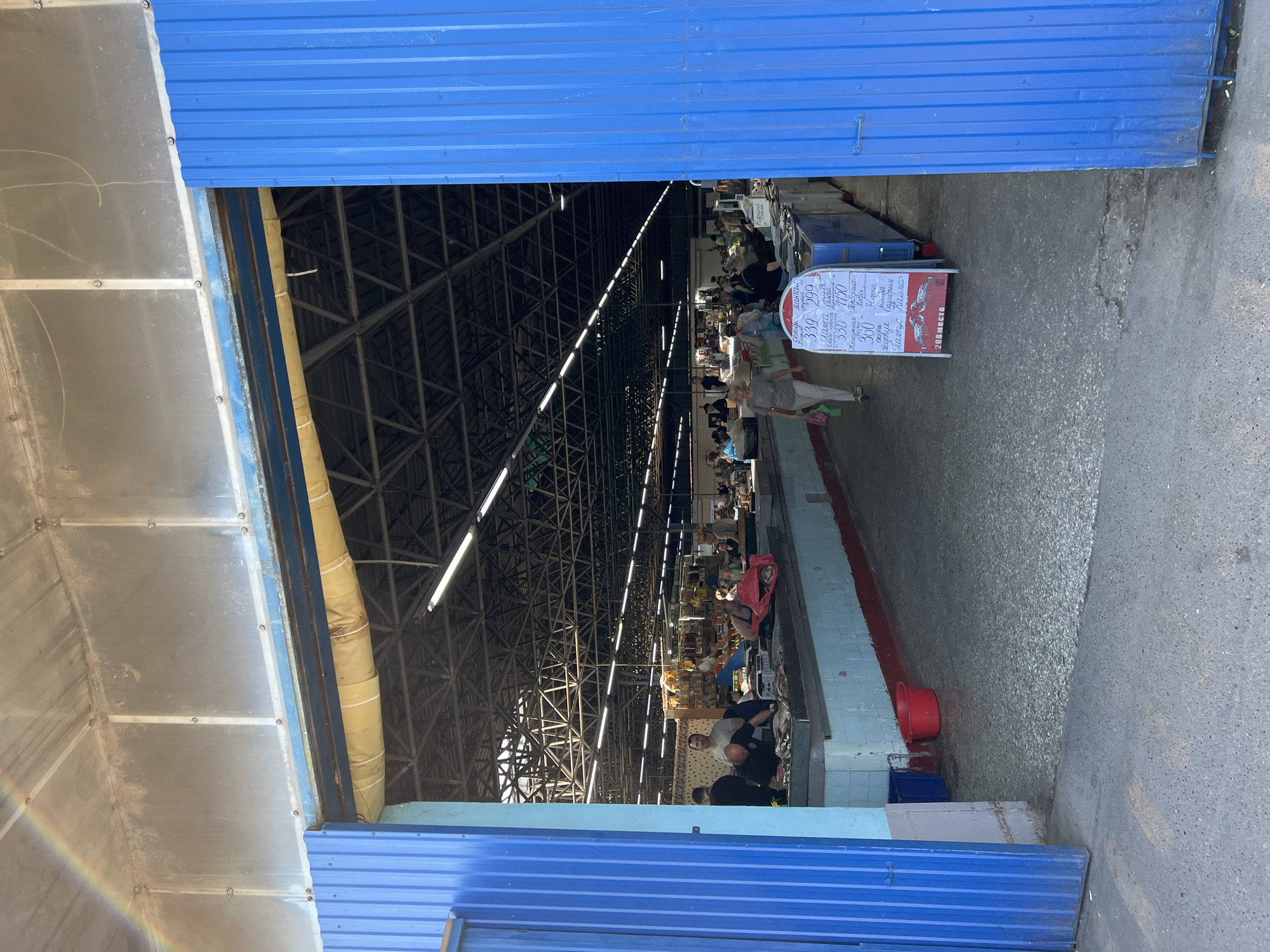 This image depicts the entrance to a covered market, identified as being in Krasnodar, Russia, based on the specific location request. The view is from just outside the market, looking inward.
The entrance is framed by two wide, open blue corrugated metal doors or walls. Above the entrance, translucent corrugated panels allow natural light into the structure, and a dirty yellow cylindrical ventilation duct is visible, running horizontally beneath the ceiling. A portion of a rainbow is visible in the upper left corner of the sky, arcing towards the top of the market structure.
The interior of the market features a high, dark, exposed metal truss ceiling structure, illuminated by numerous fluorescent strip lights. Tiled counters, predominantly light blue and white with a red base stripe, line both sides of a central aisle. On the left side, several men are gathered around a display of fish on a counter; one man in a black shirt actively handles fish, while another in a light grey T-shirt stands behind him.
Further into the market, additional people, including shoppers and vendors, are visible, though their specific activities are less distinct due to distance. A woman in light-colored attire carrying a green bag walks away from the camera in the central aisle. The market floor is a grey, speckled concrete or tiled surface, appearing damp in areas near the counters.
A prominent white A-frame sandwich board sign stands in the right foreground of the market interior. The sign displays prices and types of fish in Cyrillic, including "Семга" (Salmon), "Горбуша" (Pink Salmon), "Палтус" (Halibut), "Окунь" (Perch), "Карп" (Carp), and "Кефаль" (Mullet), with corresponding prices such as "339-299", "330-1150", and "360" (likely in Rubles). The bottom of the sign indicates "208 место" (208th spot/stall). Red plastic bags are visible on some counters, consistent with a market environment. Outside the entrance on the left, a red plastic basin and a blue plastic bin rest against the tiled base of the market wall.