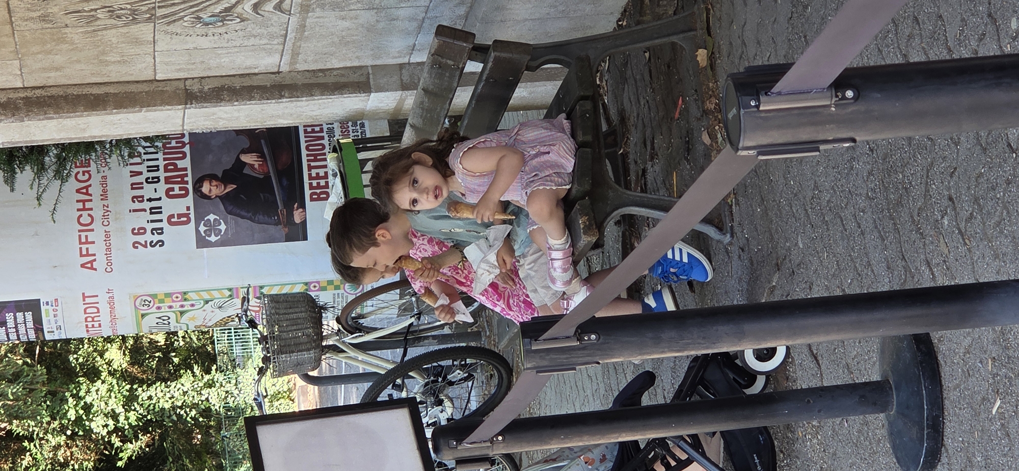 This media file depicts two young children sitting on a park bench and eating ice cream. The boy is wearing a pink and white patterned shirt, while the girl is wearing a pink striped dress. They are both holding ice cream cones. Behind them, a poster advertises an event featuring "G. CAPUCI" on "26 Janvie" at "Saint-Guil". Another poster mentions "BEETHOVEI". The scene appears to be outdoors, with trees and cobblestone paving visible. A bicycle with a wicker basket is parked next to the bench. The overall impression is one of a casual, pleasant day in a public space. The lighting suggests it is daytime. The children seem content and engaged in their treat. The presence of the posters and the park bench suggests a public area, possibly near a venue or event space. The location is indicated as Strasbourg, France.