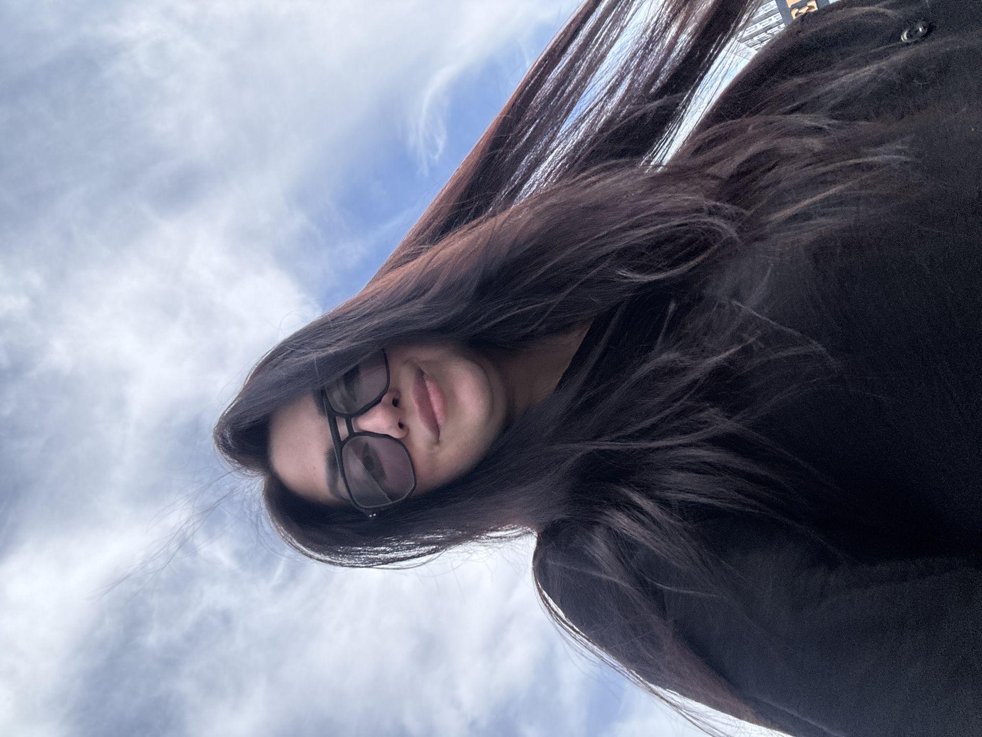 A woman with long, dark hair and glasses is looking down at the camera. The sky is visible in the background.