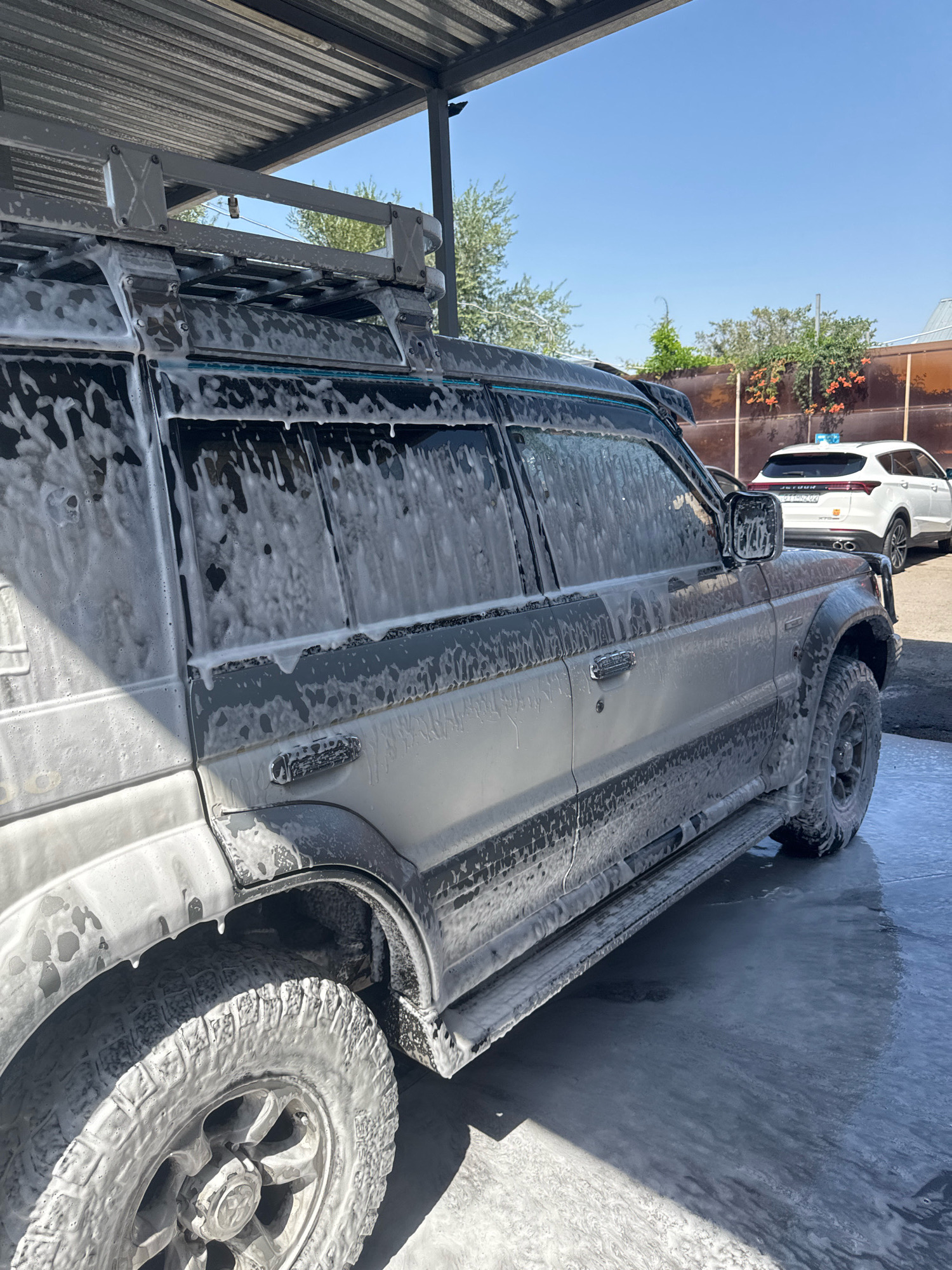 A dark-colored SUV, covered in white foam from a car wash, is parked under a metal awning. The vehicle is angled to the right, with its front end obscured. The foam is particularly thick on the roof rack, windows, and tires. The ground around the vehicle is wet and covered in suds, indicating an active car wash. In the background, a white SUV with a blue light on its roof is visible parked behind the first vehicle. The scene appears to be an outdoor car wash facility with a clear blue sky overhead and trees and a fence in the distance. The location is identified as Almaty, Kazakhstan.
