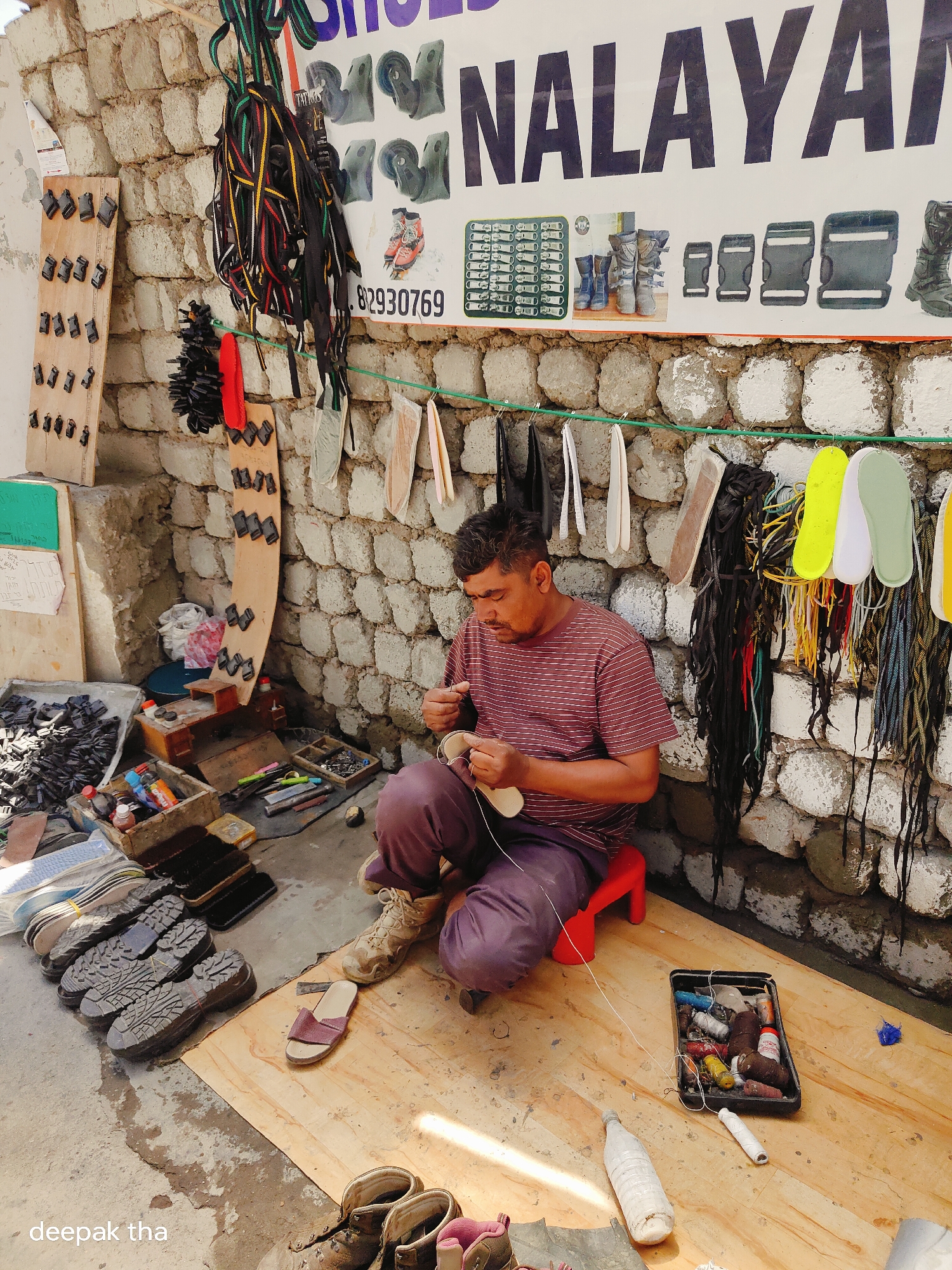 In Leh, India, a cobbler is working on footwear outdoors. He is seated on a red stool and is engaged in sewing a sandal. The surrounding environment is cluttered with various shoe-making materials, including stacks of shoe soles, rolls of straps, and sewing supplies. The backdrop consists of a stone wall displaying promotional materials for "HIMALAYAN SHOES" and other shoe accessories.