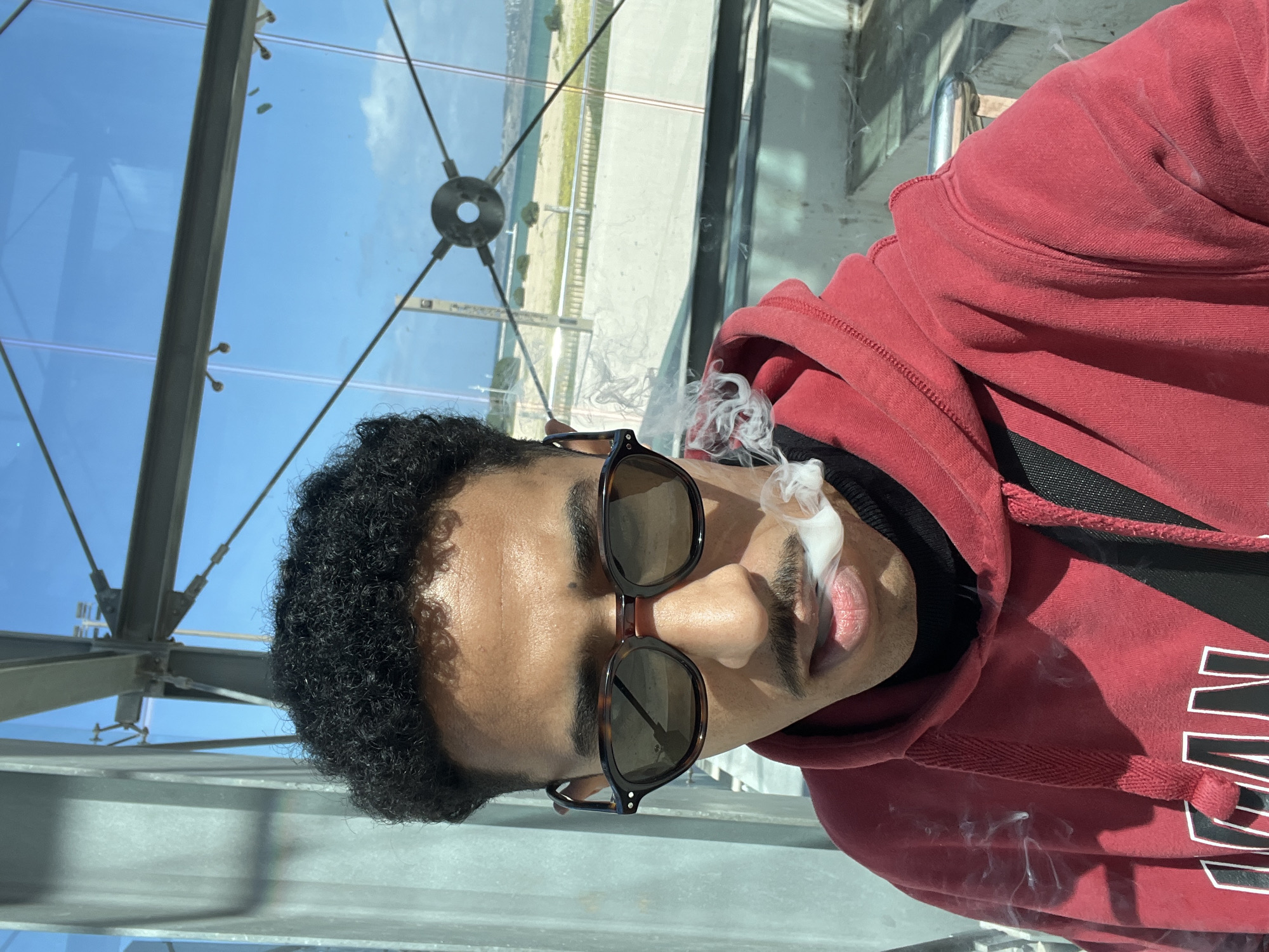 A young male subject with dark, curly hair and a distinct mustache is depicted in a close-up image, exhaling a plume of white vapor or smoke from his mouth. He wears tortoiseshell-patterned sunglasses, a red hooded sweatshirt over a black turtleneck, and a black strap of a bag diagonally across his chest. The subject is situated within a modern, glass-enclosed structure, likely an elevated walkway or viewing area, as evidenced by visible grey metal support beams, cables, and connectors for the large glass panels.
Through the transparent panels, a bright, clear blue sky with minimal white clouds is visible. The background reveals a coastal environment, featuring a light-colored terrain, possibly sand or dry vegetation, a concrete or paved walkway, and a metallic railing. Beyond these elements, a significant body of water, consistent with the sea, extends to a distant coastline or landmass. The architectural style and coastal view are consistent with a location like the port or a modern waterfront development in Tanger, Morocco.