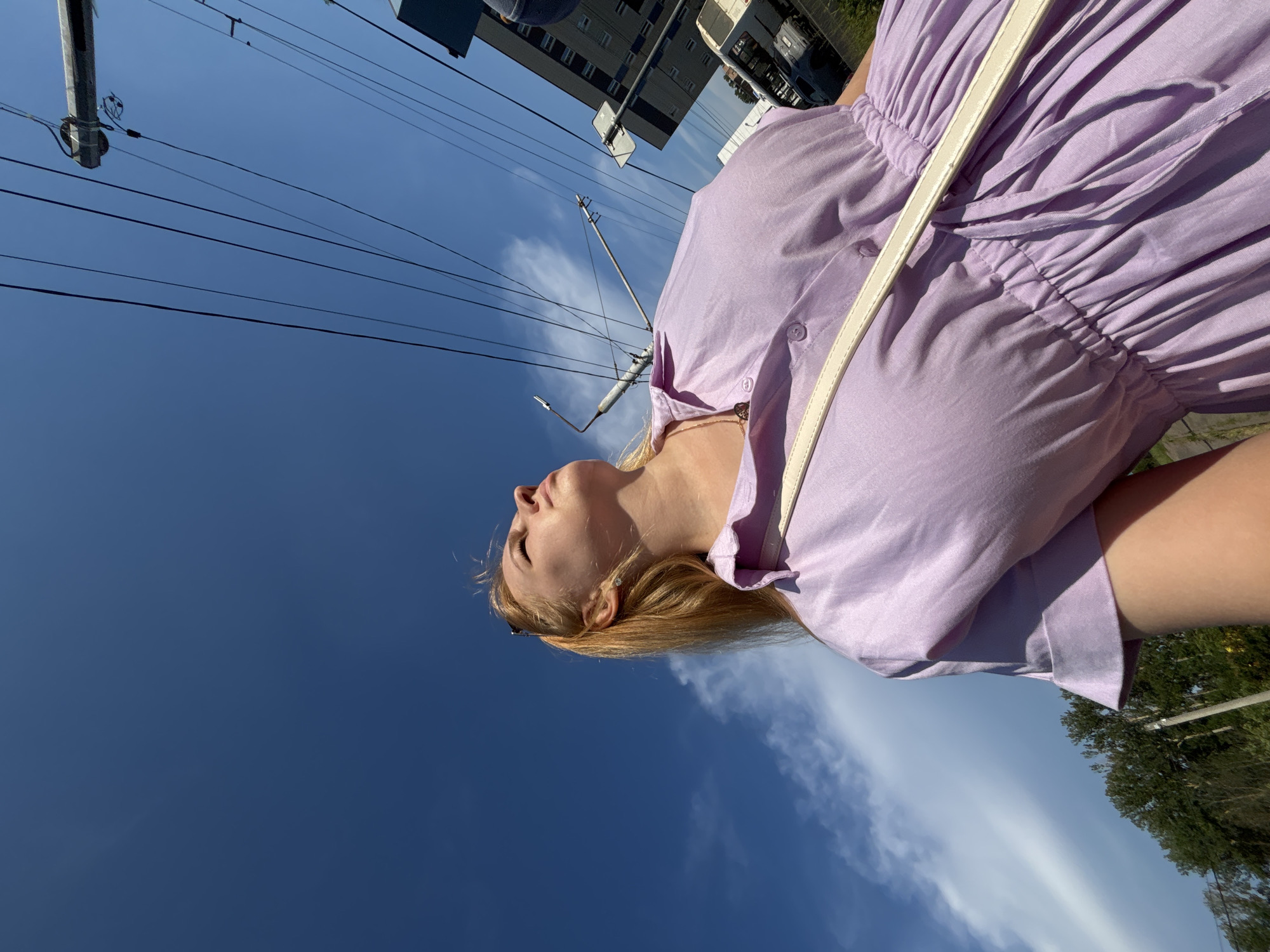 The image shows a young woman from the chest up, looking upwards against a clear blue sky. She has blonde hair and is wearing a light purple dress with short sleeves and a collared neckline. A white strap from a bag is visible across her chest. In the background, numerous power lines crisscross the sky, with a utility pole visible on the left. To the right, a tall building with multiple windows and a bus are partially visible, suggesting an urban environment. The low-angle shot emphasizes the vastness of the sky and the woman's upward gaze. The scene appears to be an outdoor public space in the city of Zavolzhye, Russia, likely during daylight hours.